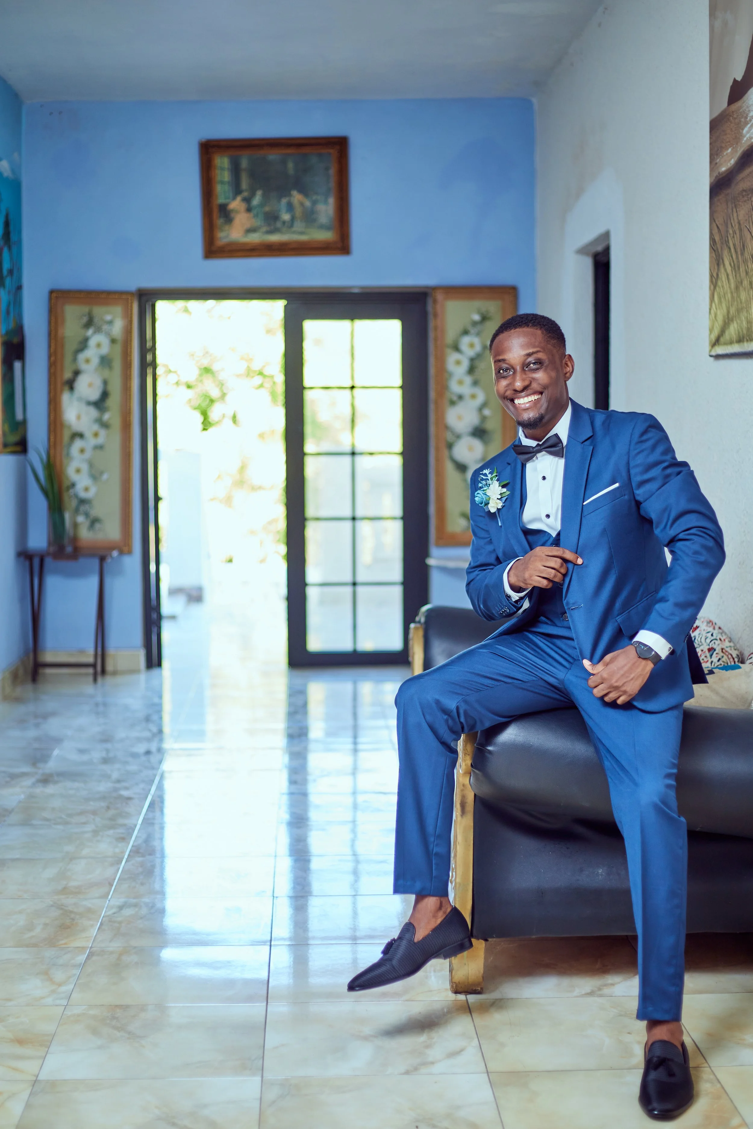 A man in a royal blue tuxedo with a black bow tie, white shirt, and matching boutonniere sitting on a black leather couch inside a well-lit room, smiling at the camera.