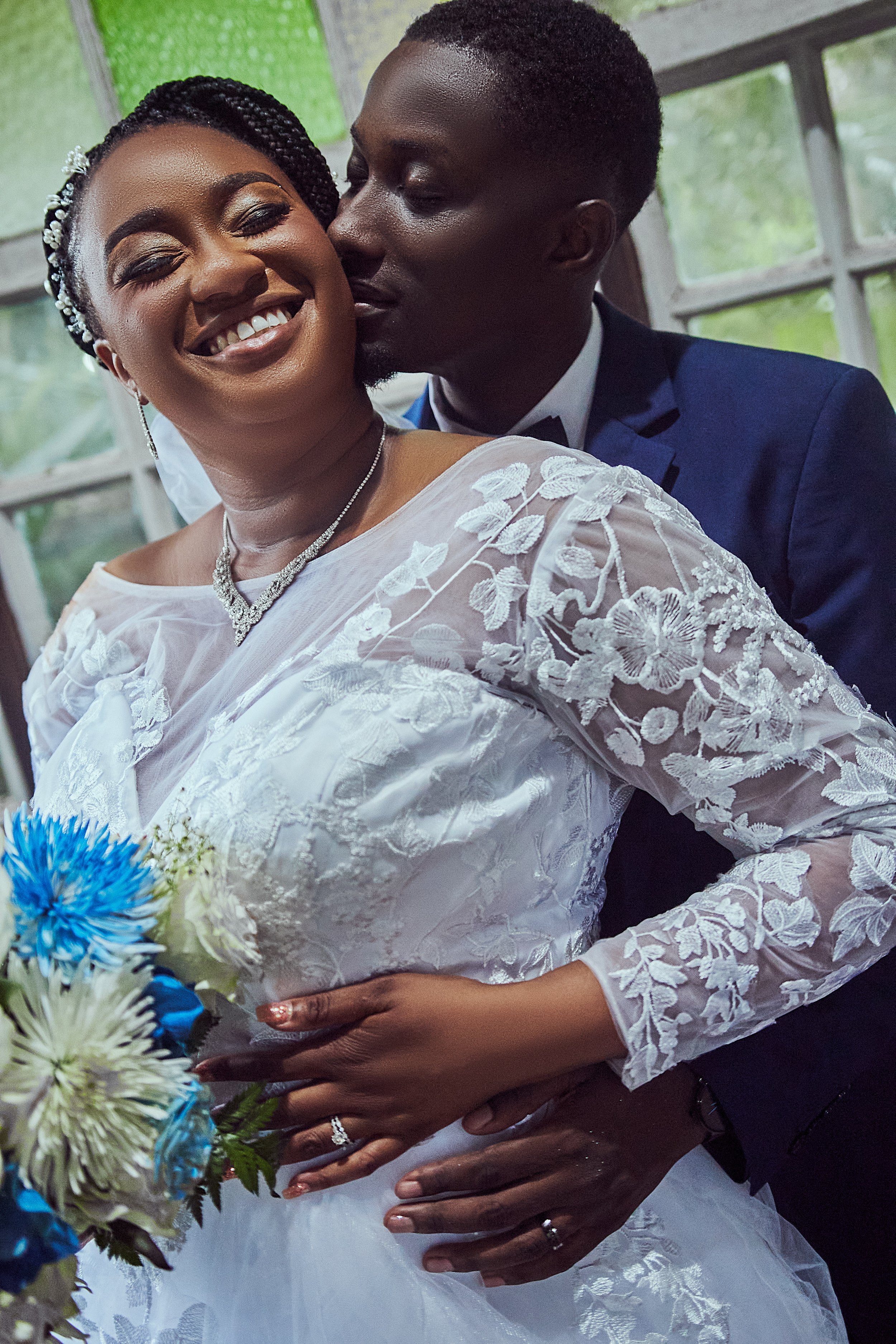 A joyful bride and groom on their wedding day, with the groom whispering into the bride's ear, inside a lit room with large windows.