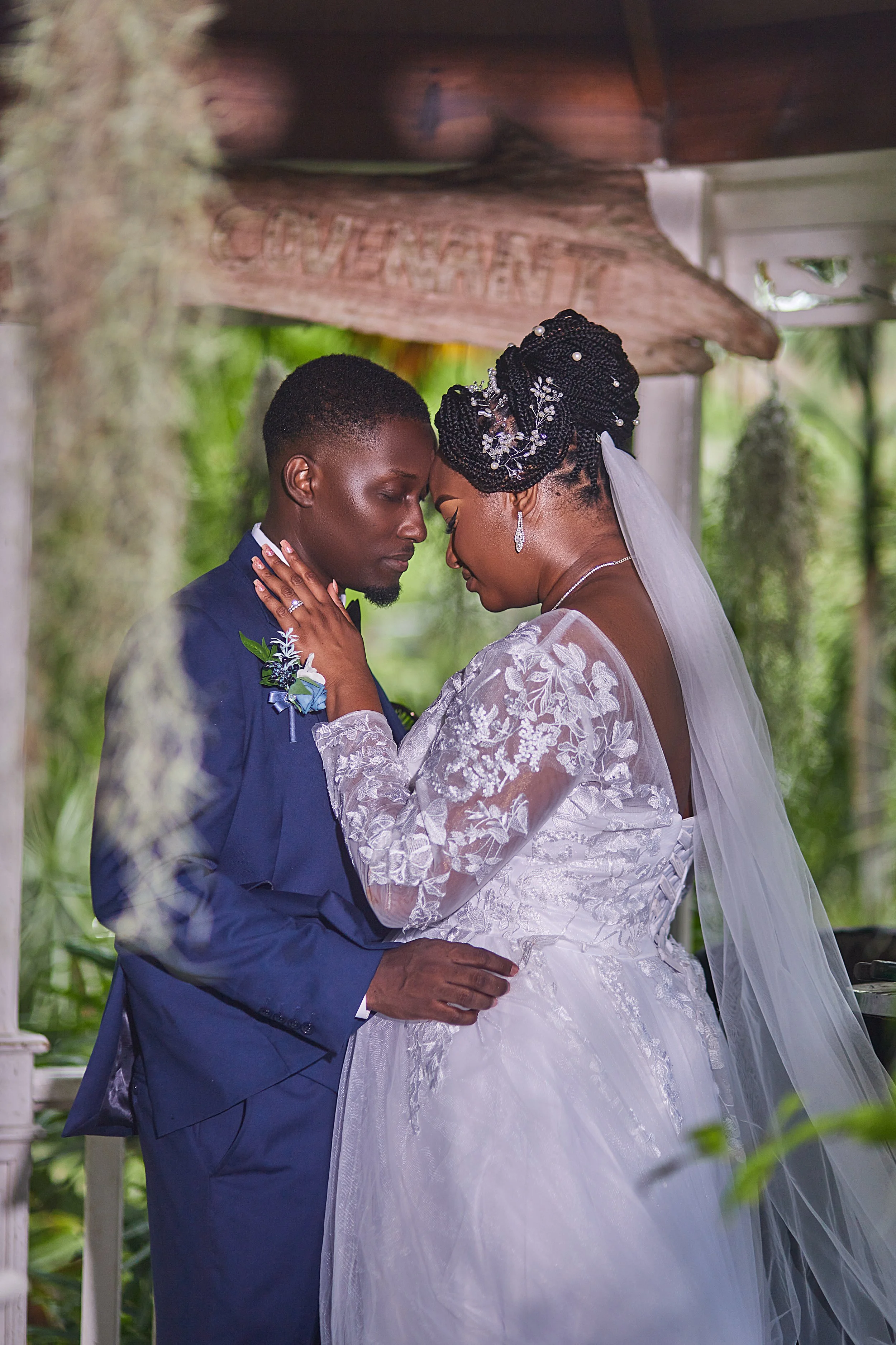 A bride and groom sharing an intimate moment during their wedding, standing close with foreheads touching, surrounded by greenery.