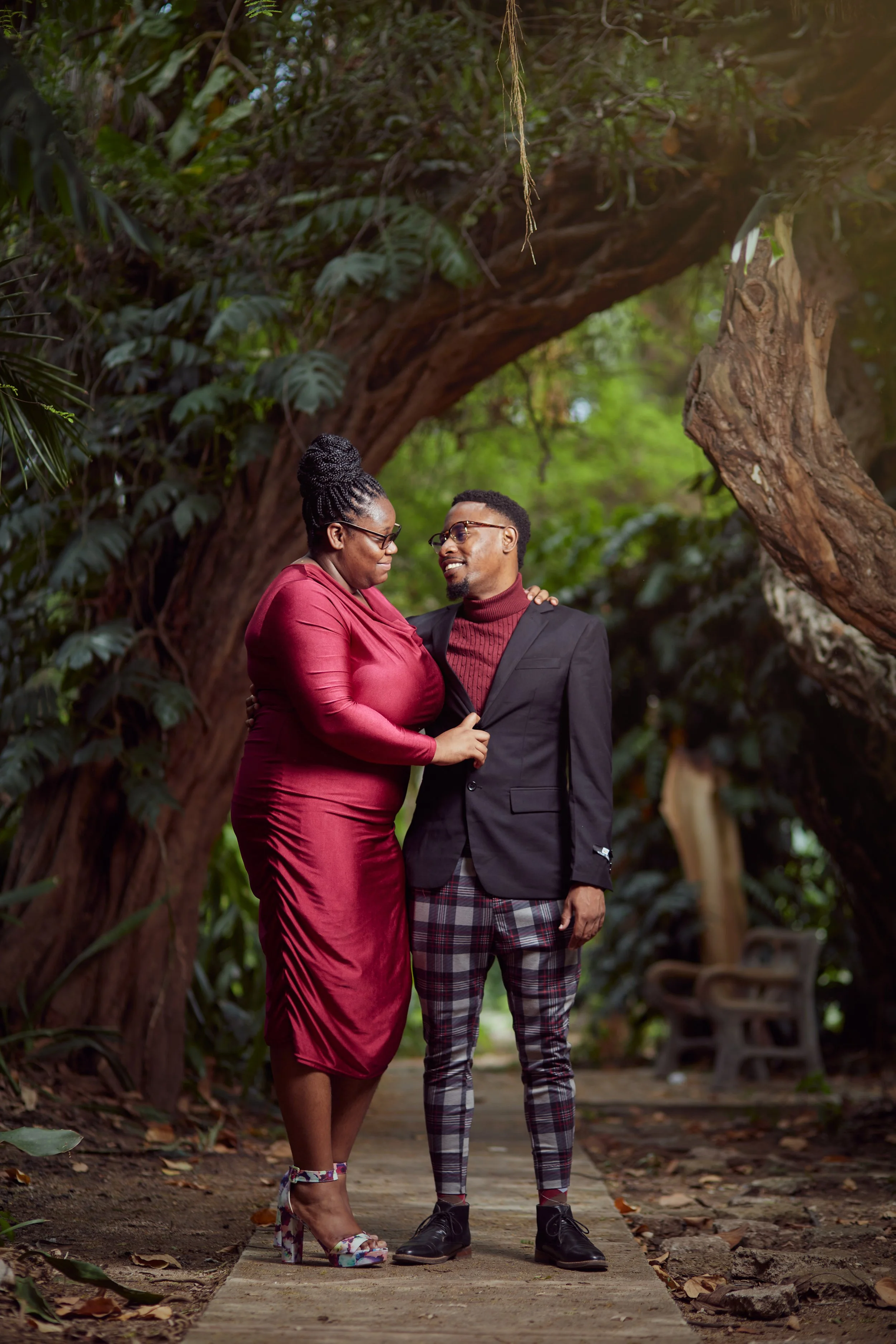 A woman and a man standing close under a large, arching tree in a lush, green park are smiling at each other.
