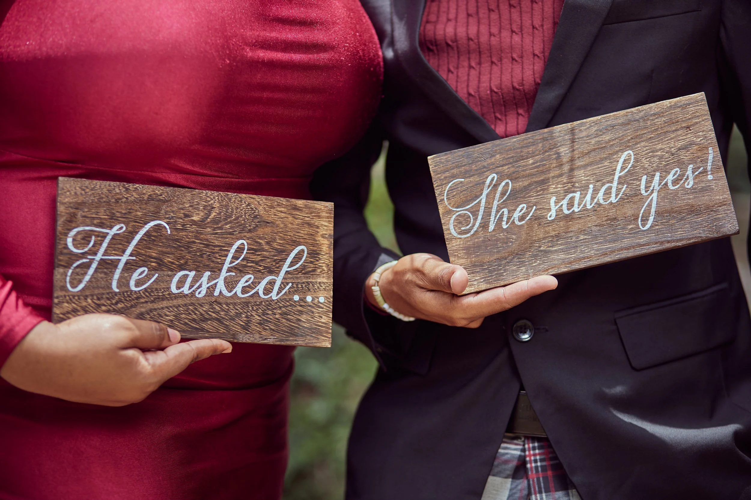 Two people holding wooden signs, with one reading "He asked..." and the other reading "She said yes!"