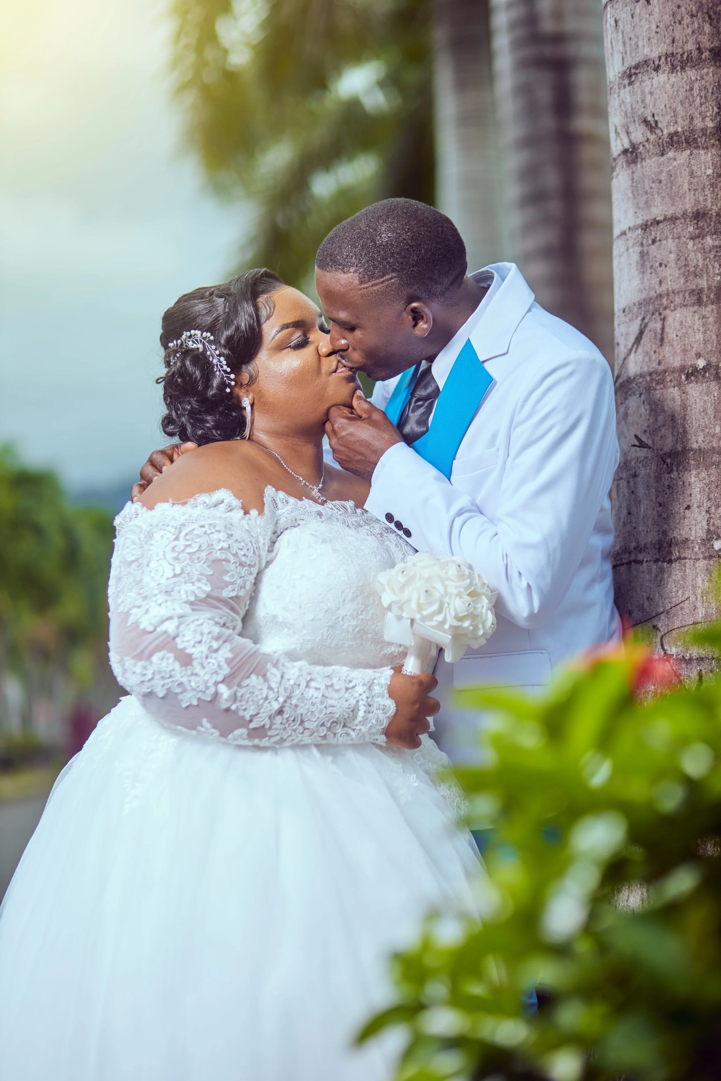 A newlywed couple sharing a kiss outdoors, the bride holding a bouquet and wearing a white lace wedding dress, and the groom in a white suit with a blue tie, leaning against a tree.