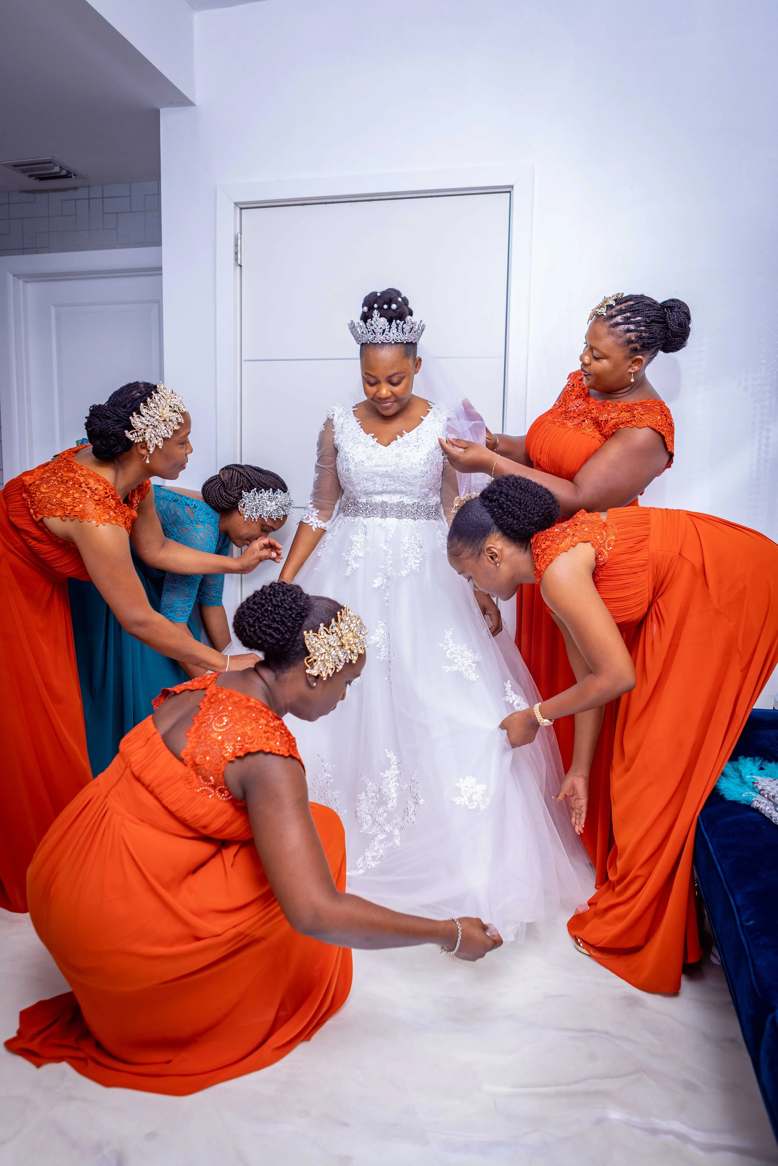 A bride in a white wedding dress with a tiara is surrounded by five women, four in orange dresses and one in a blue dress, helping her prepare for her wedding.