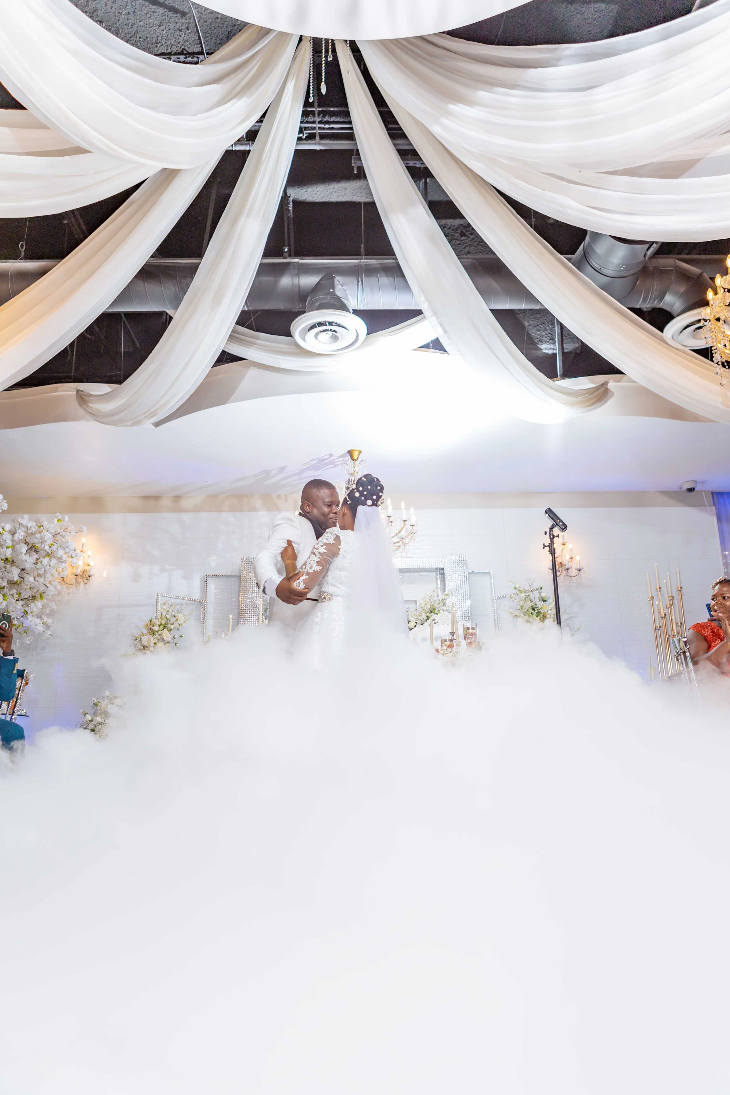 A bride and groom share their first dance amidst a foggy effect at the wedding reception, surrounded by elegant drapery, floral arrangements, and soft lighting.
