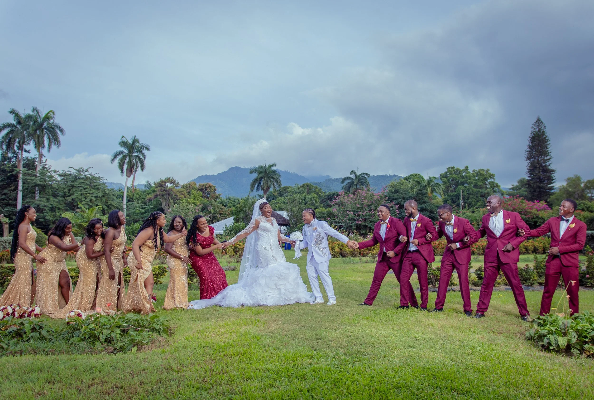 A wedding party standing in a line outdoors on a grassy field, holding hands. The bride and groom are in the center, with the bride wearing a white wedding dress and veil and the groom in a white suit. Bridesmaids in gold dresses are on one side, and