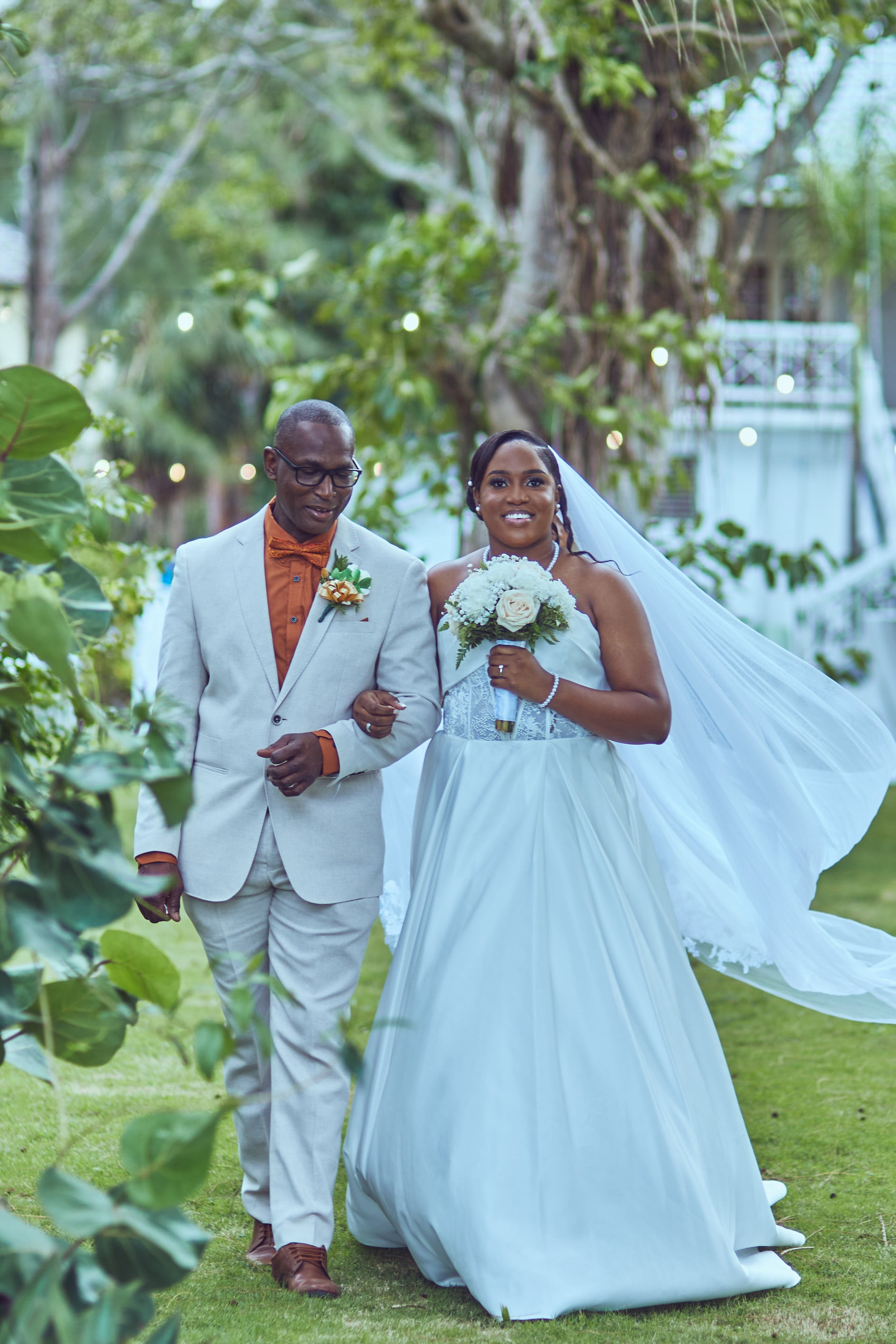 A bride in a white wedding dress holding a bouquet of white roses, walking arm-in-arm with a man in a light gray suit and orange shirt and bow tie, outdoors with lush green trees.