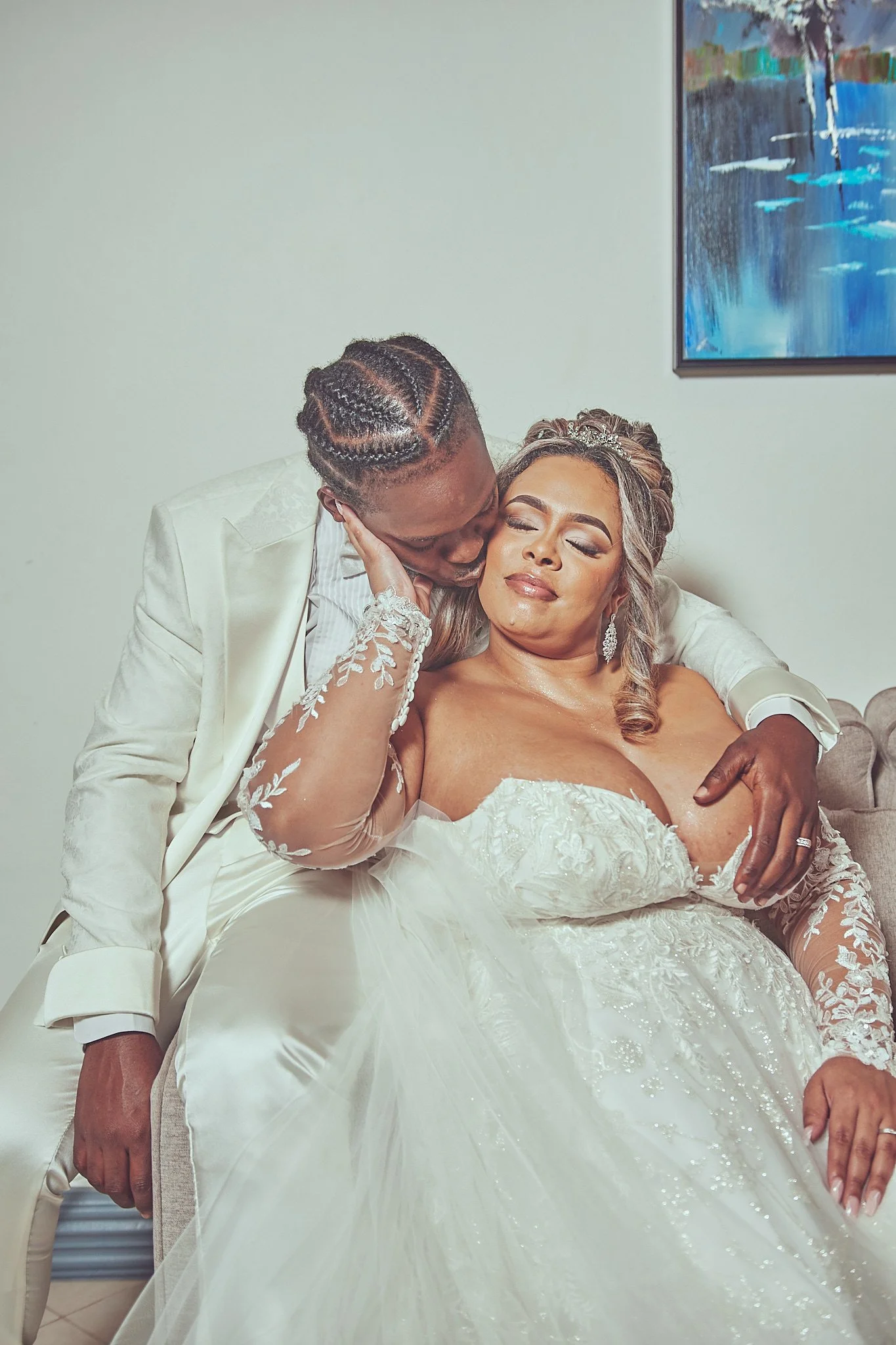 A bride and groom embracing on their wedding day. The groom is kissing the bride on her cheek while she is sitting on a couch, wearing a white wedding gown and jewelry, with her eyes closed.