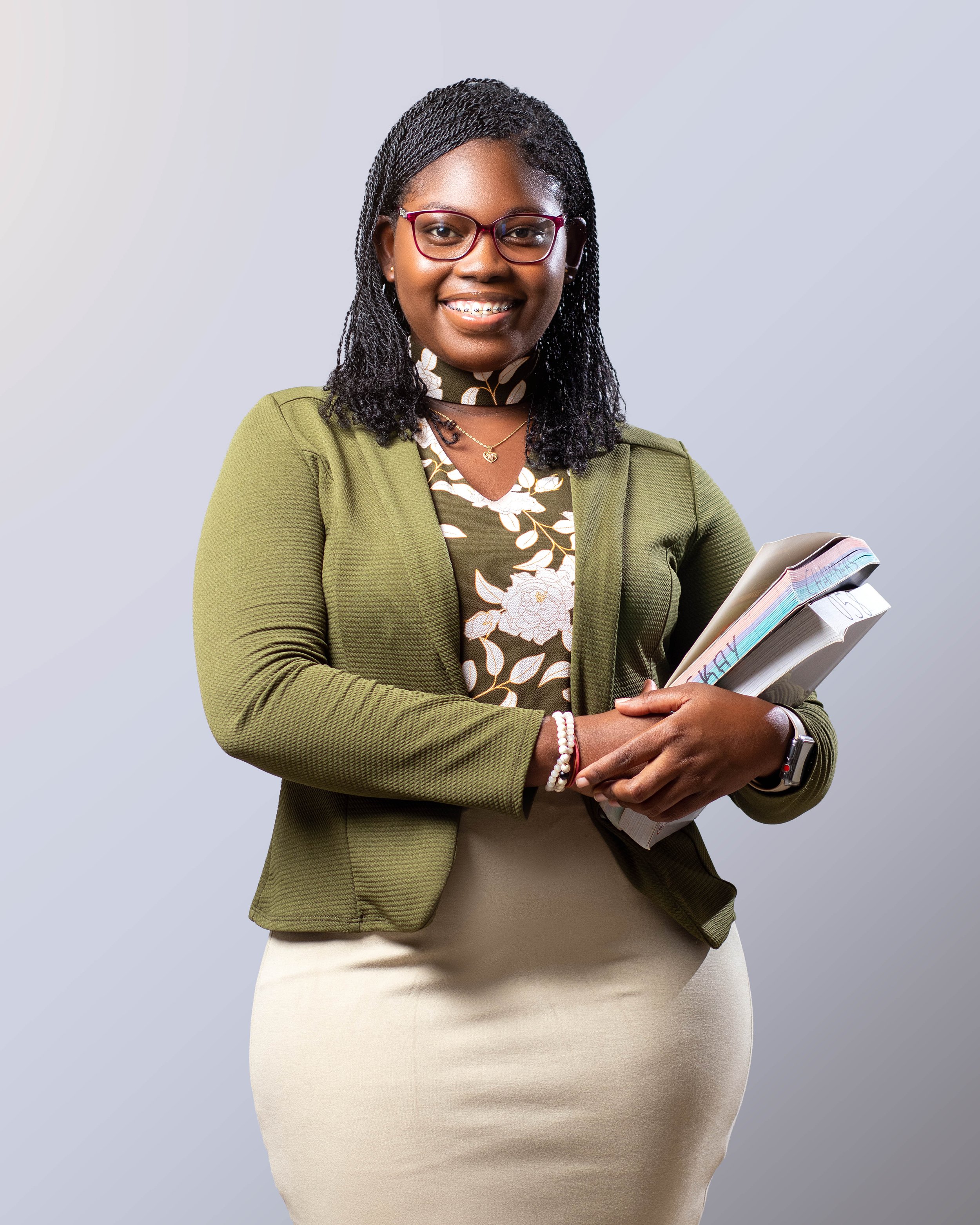 A woman smiling, holding folders and documents, wearing glasses, a green blazer, a floral blouse, and a keyboard in her hand.