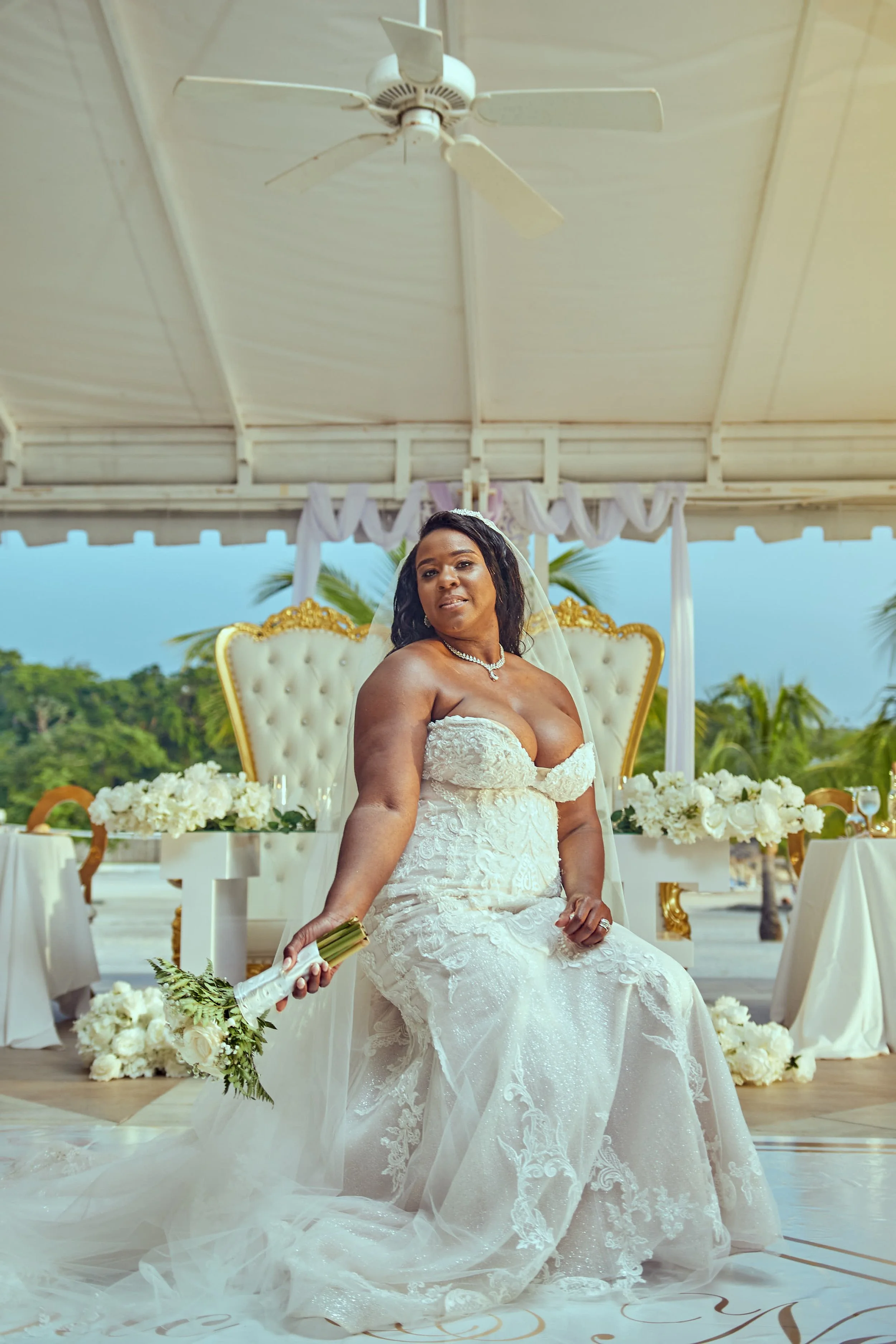 A bride in a white wedding dress sitting on a throne-like seat, holding a bouquet, at her wedding reception outdoor under a canopy with floral decorations and palm trees in the background.