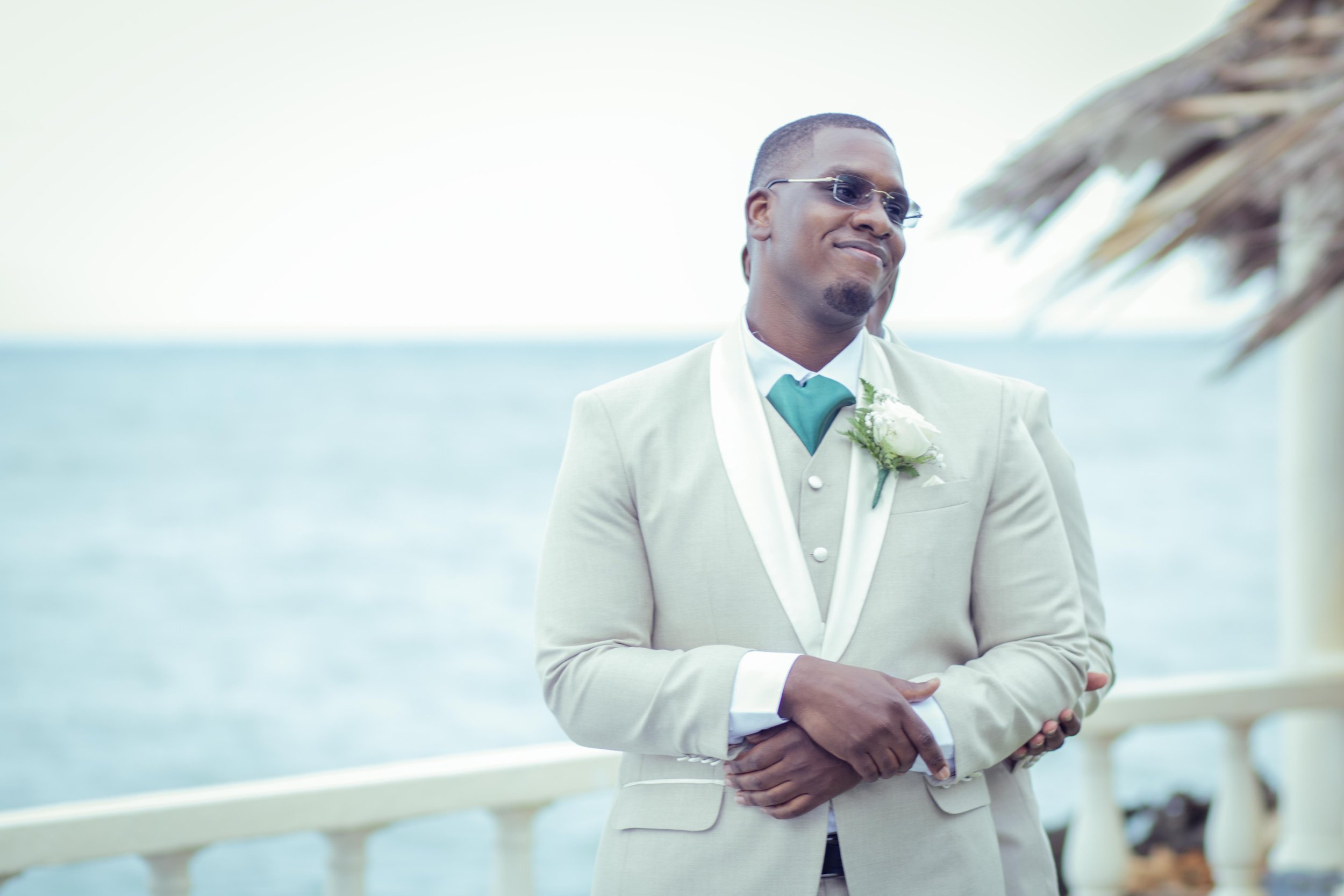 A man in a light suit with sunglasses and a flower boutonniere standing outdoors near the water with a thatched roof in the background.