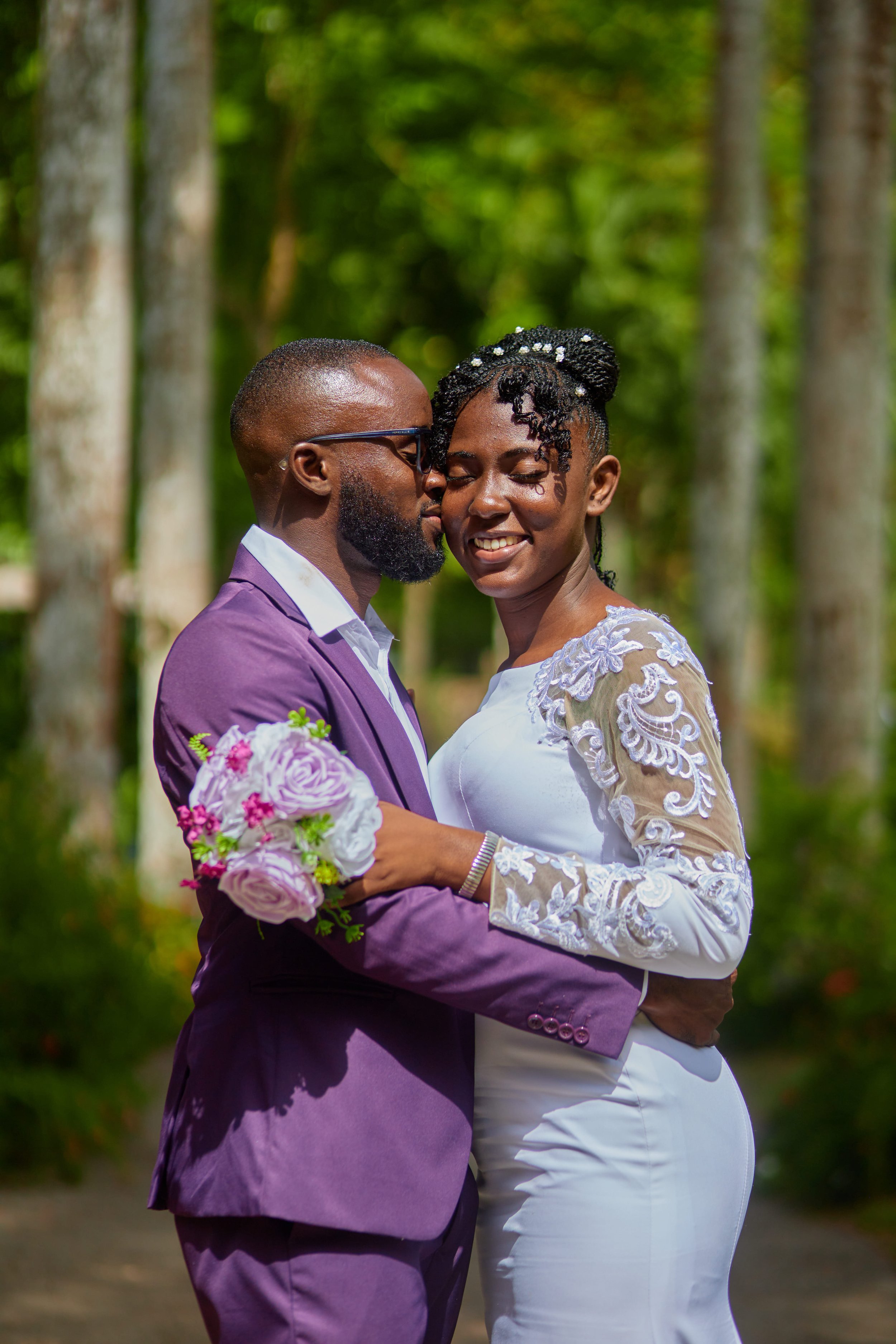 A couple dressed in wedding attire sharing a tender moment outdoors, surrounded by green trees, with the groom holding a bouquet of pink and purple flowers.