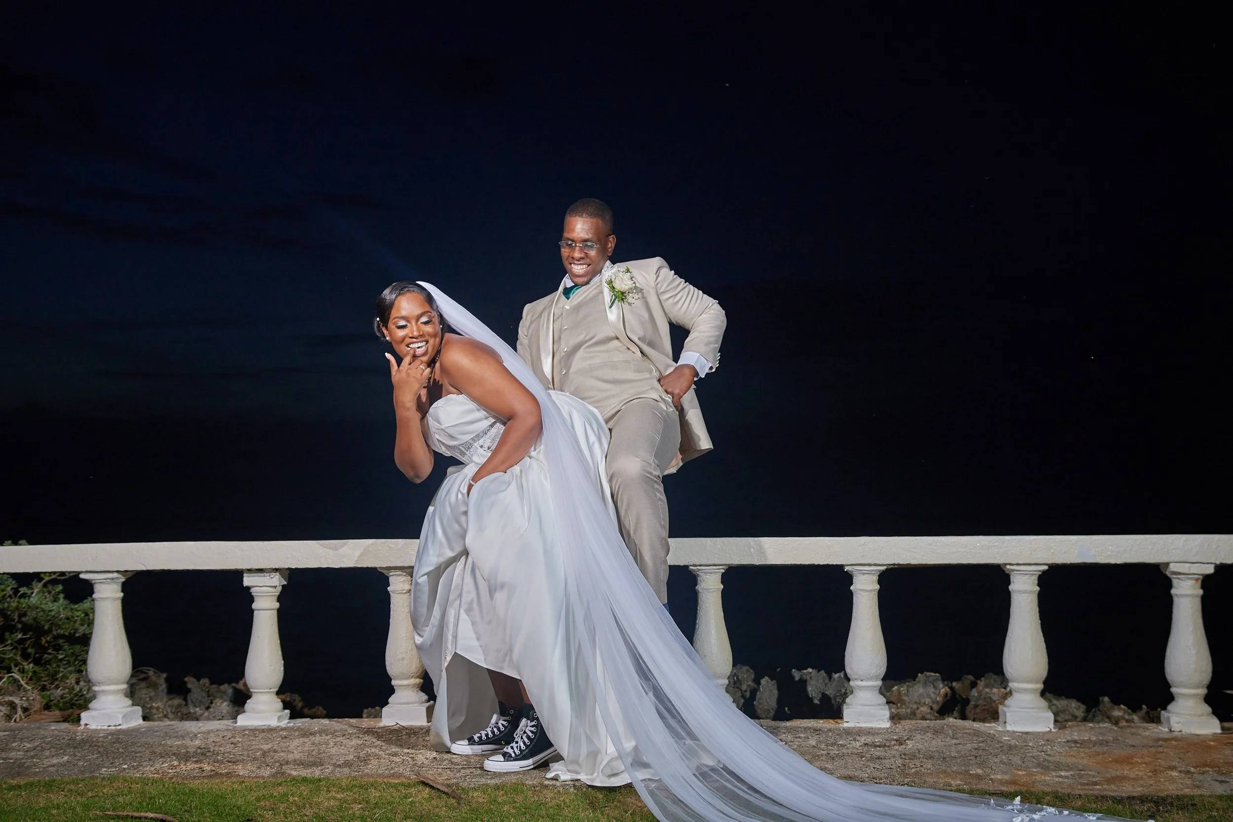 A newlywed couple at night, with the bride wearing a white wedding gown and veil, and the groom in a light-colored suit, standing outdoors near a white railing by the water, both smiling and posing playfully.