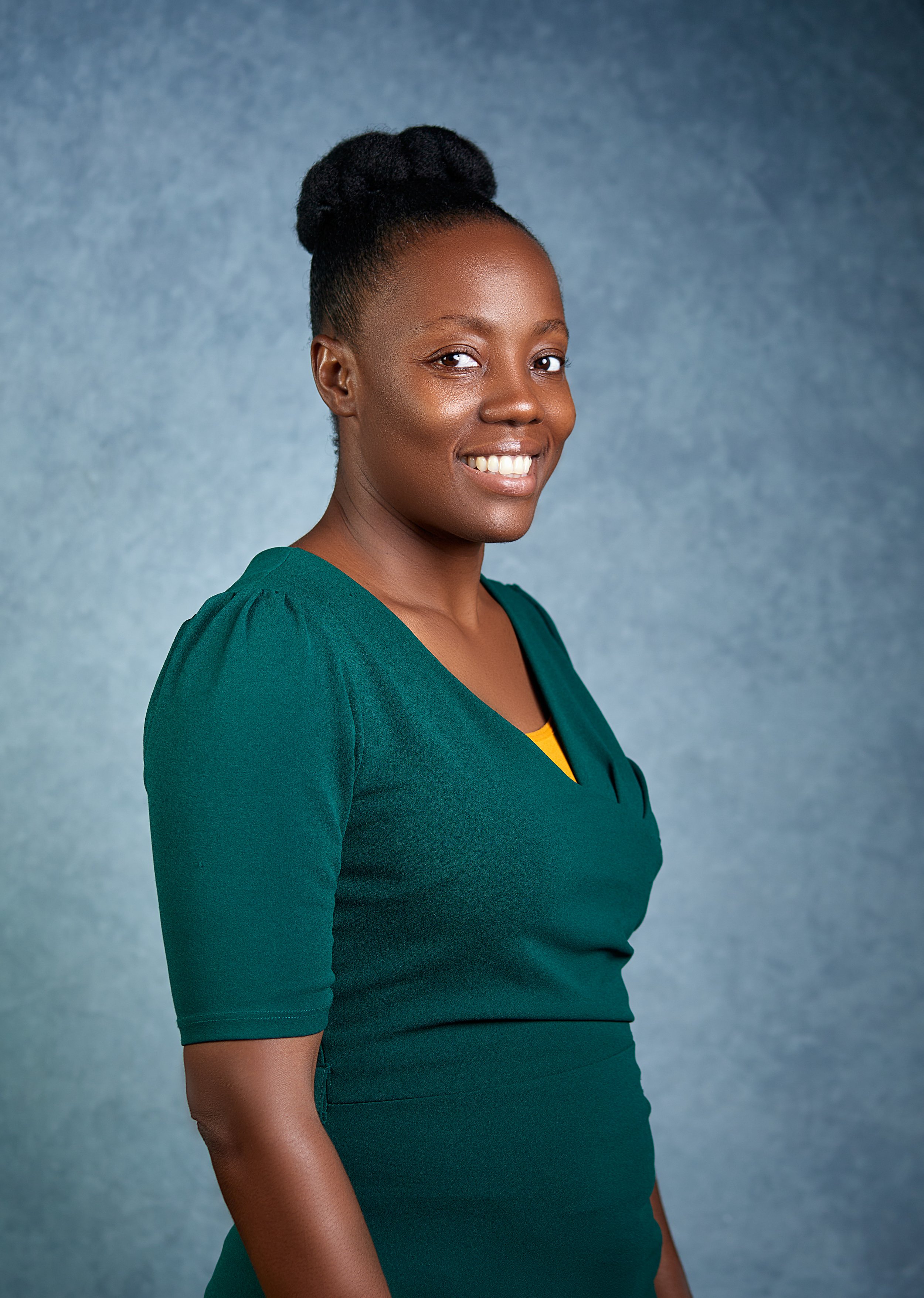 A woman with dark hair styled in an updo, wearing a dark teal dress with a yellow top underneath, smiling against a textured blue background.