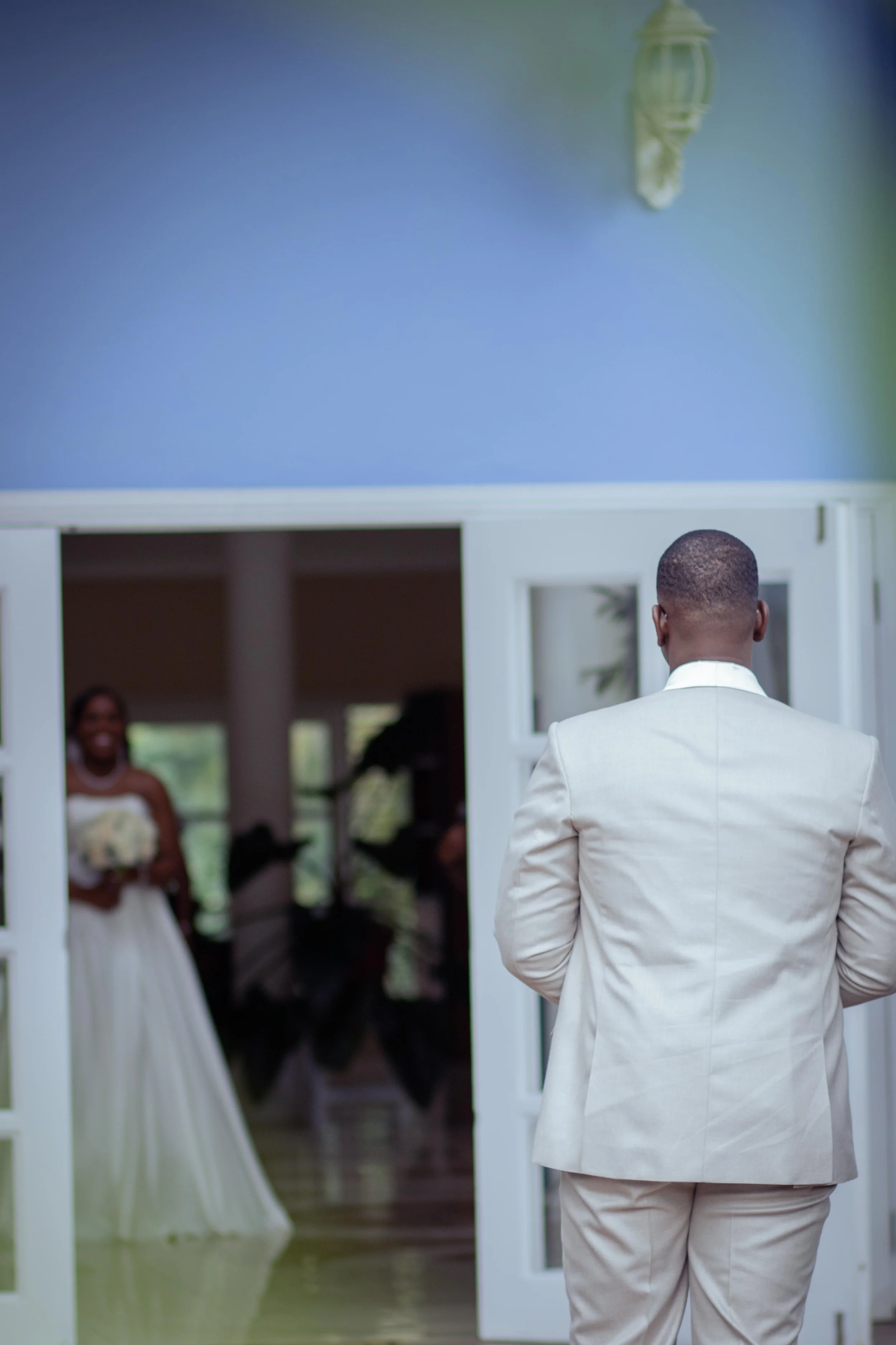 Man in a white suit watching a woman in a white dress holding a bouquet. They are indoors near glass doors, with a woman smiling in the background.