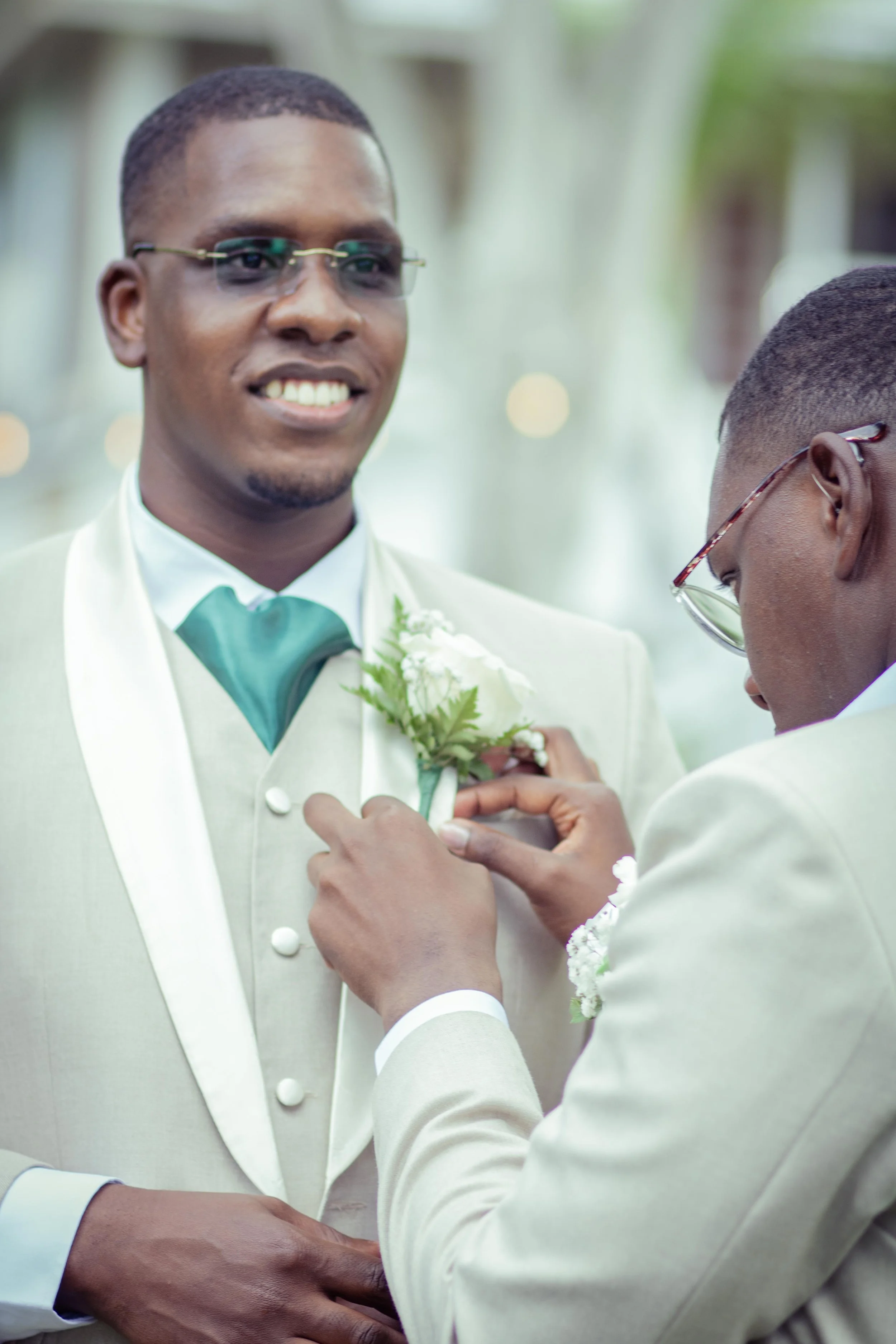 Two men in cream suits at a wedding, one is pinning a boutonniere on the other's jacket.