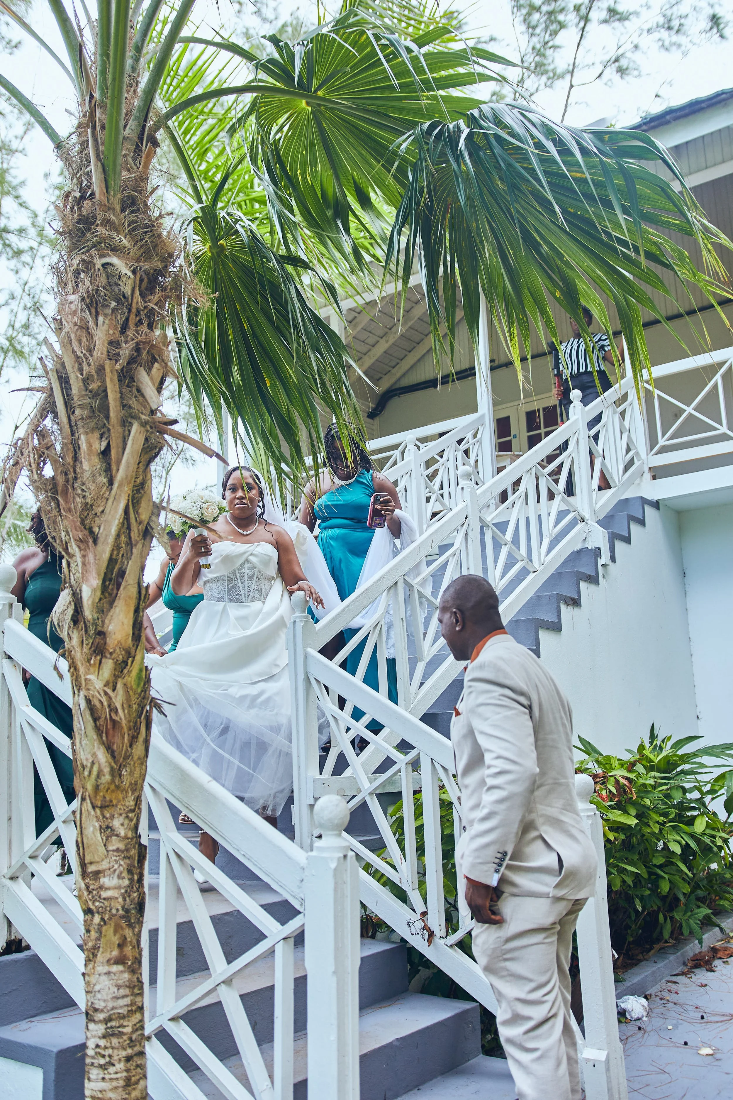 A woman in a white wedding dress sitting on white stairs outside a house, with other women around her, and a man in a cream suit nearby, under a tree.