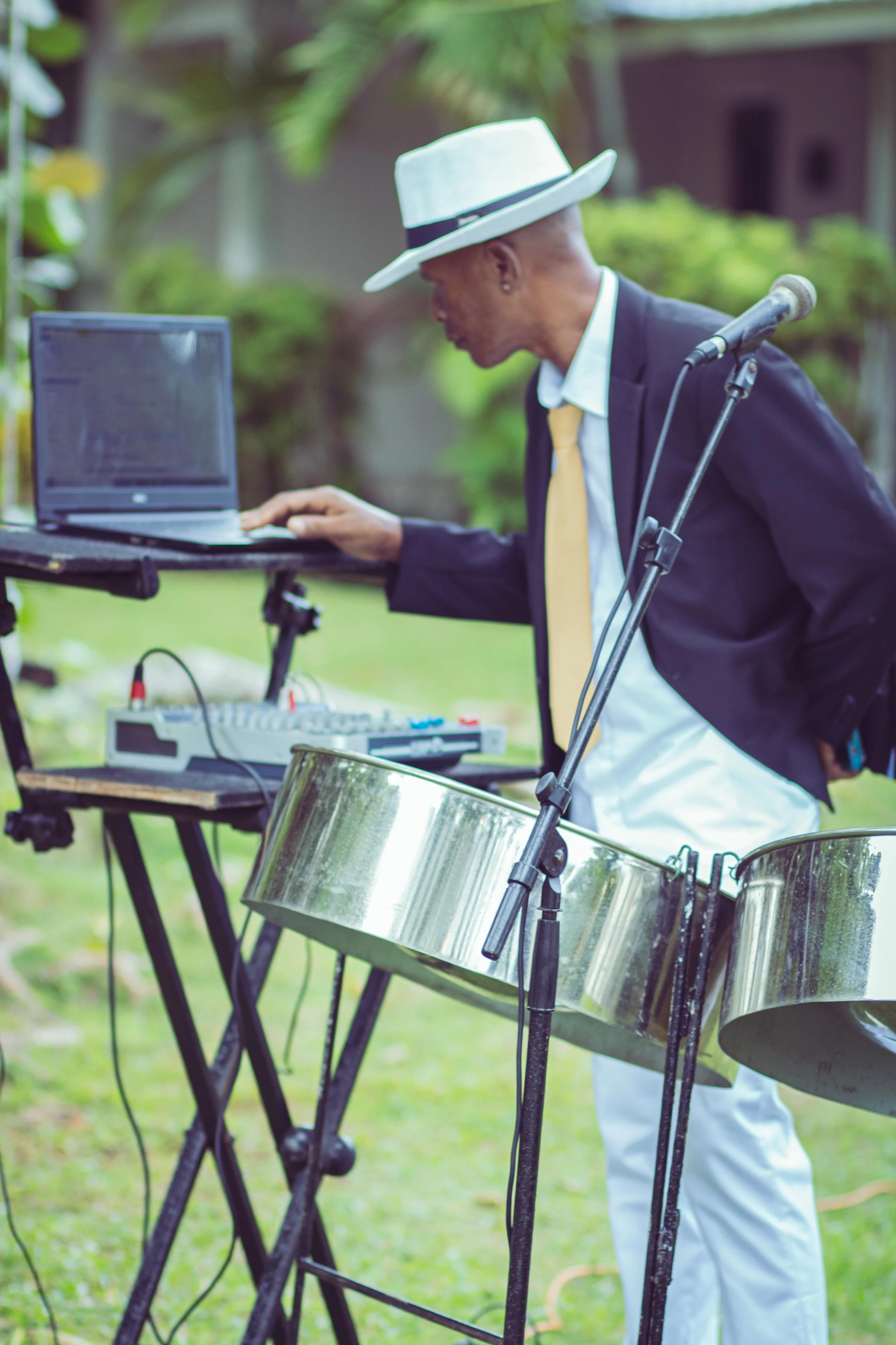 A man wearing a white hat, black suit jacket, white shirt, and yellow tie stands outdoors. He is operating a laptop and music equipment, including a steel drum and a small mixer, with a microphone in front of him.