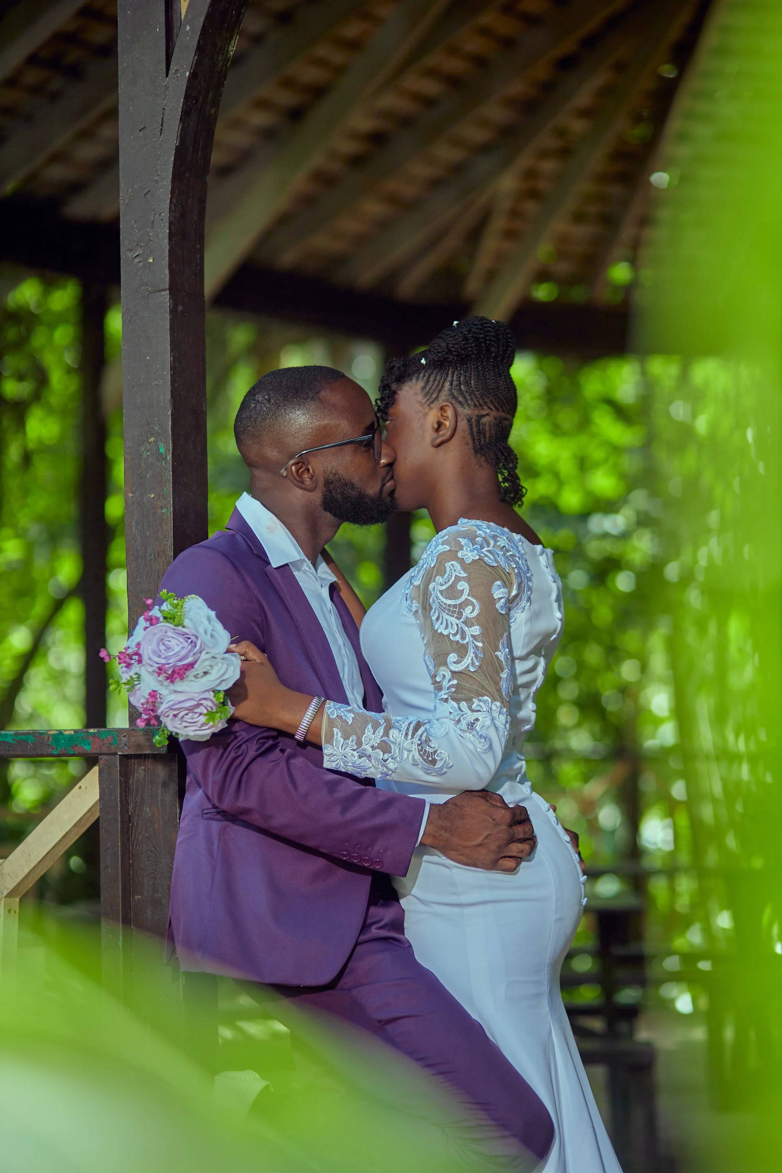 A newlywed couple sharing a kiss in an outdoor setting with lush greenery, a wooden gazebo, and sunlight filtering through the trees.