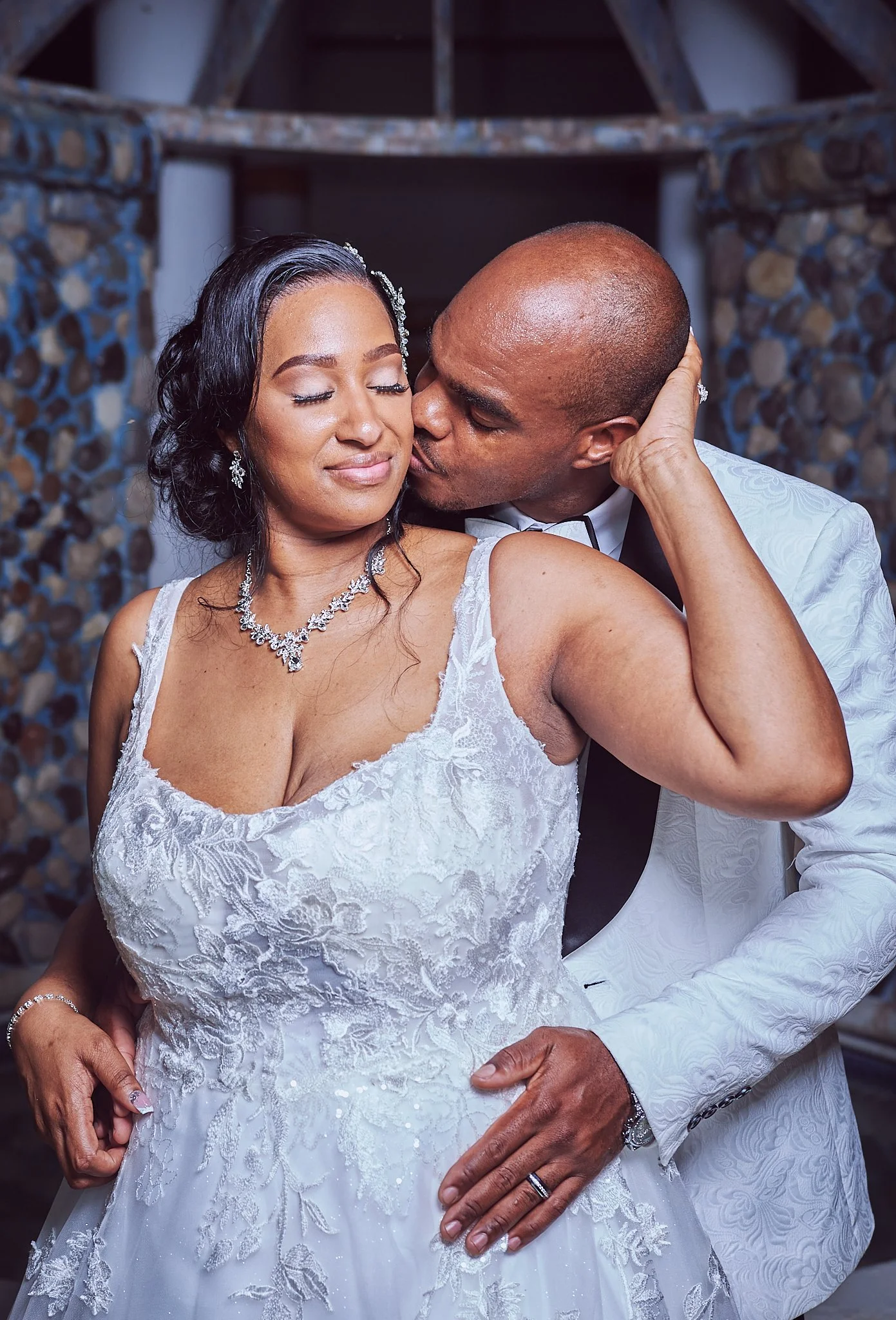 A bride and groom share an intimate moment on their wedding day, with the groom kissing the bride's cheek and holding her waist. The bride is wearing a detailed white wedding gown with lace and jewelry, while the groom is in a white tuxedo with a bla