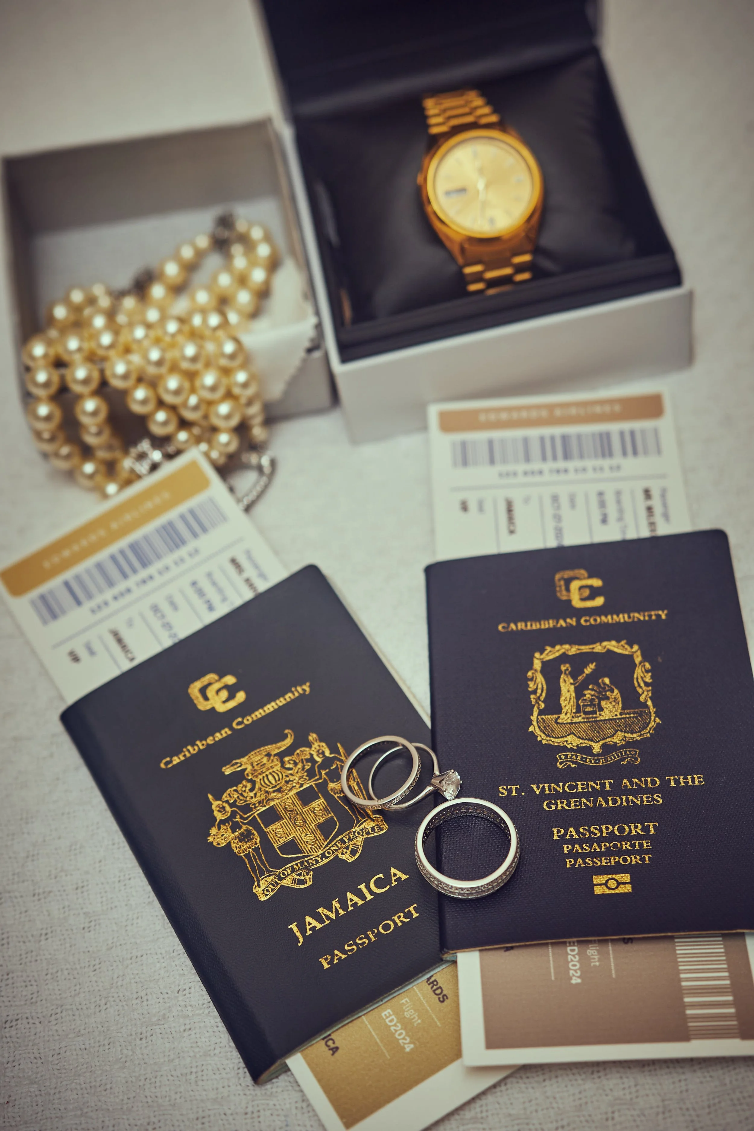 Travel essentials including two passports from Jamaica and St. Vincent and the Grenadines, wedding rings, a gold wristwatch in a box, a pearl necklace, and boarding passes on a light-colored surface.