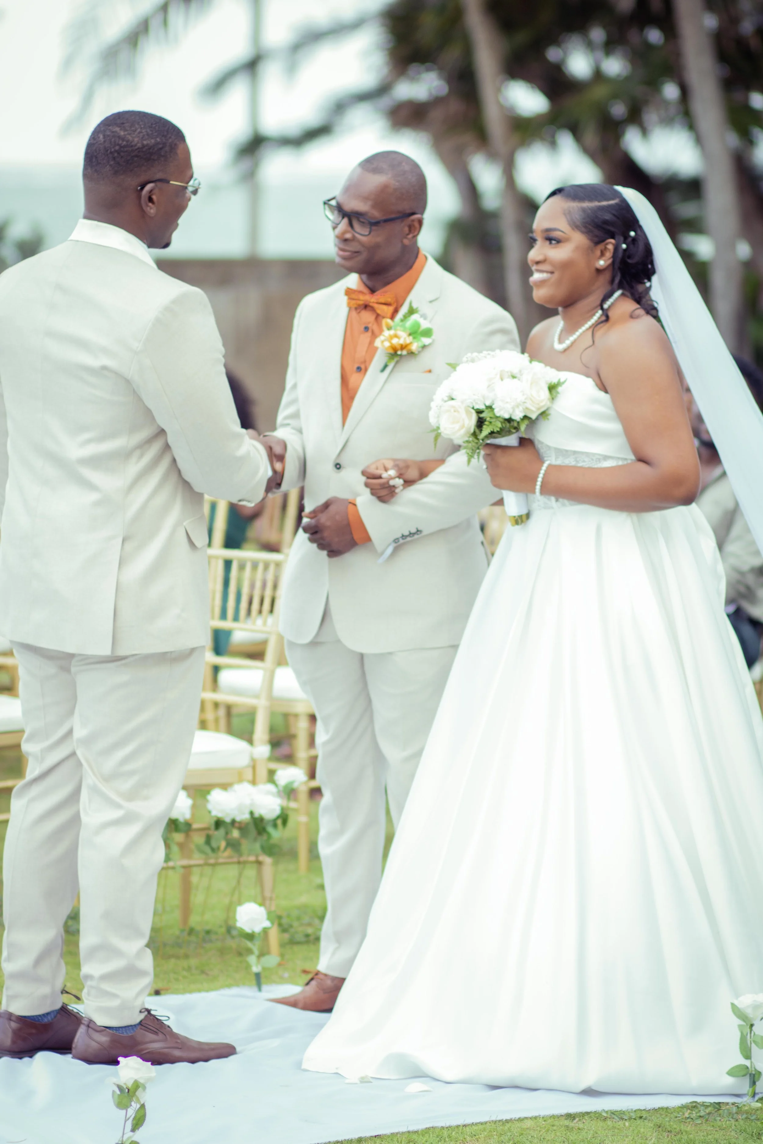 A wedding ceremony taking place outdoors with a bride in a white gown holding a bouquet, a groom in a beige suit, and an officiant in a light gray suit shaking hands. There are chairs and floral decorations in the background.
