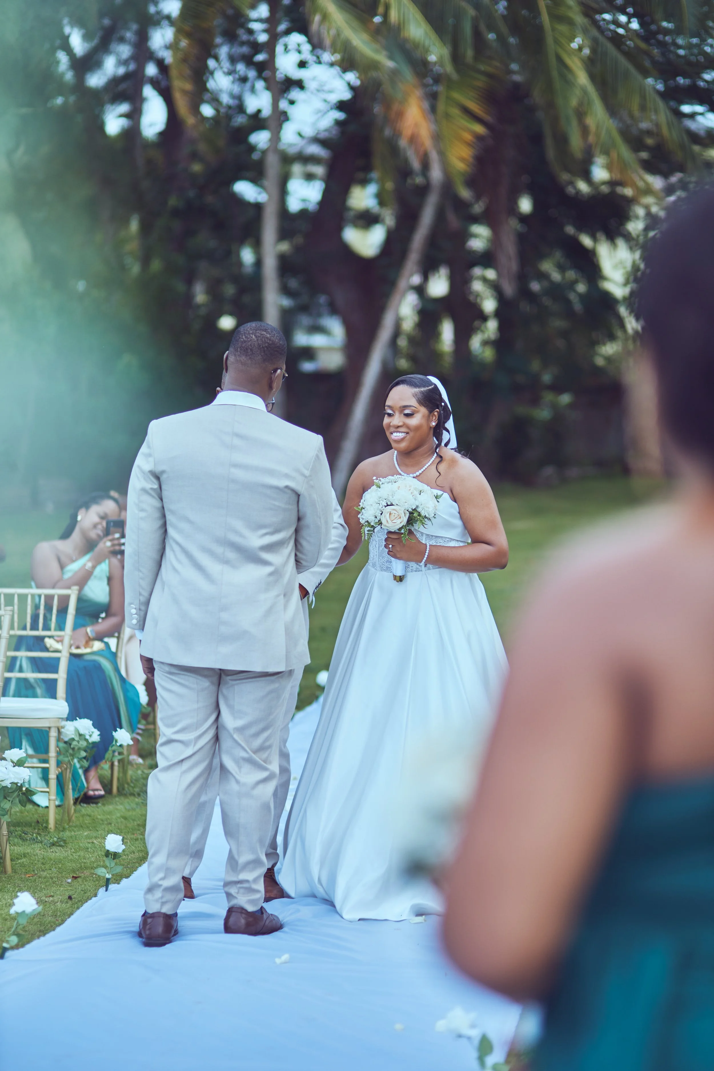 A bride and groom exchanging vows outdoors during a wedding ceremony, with guests seated nearby.