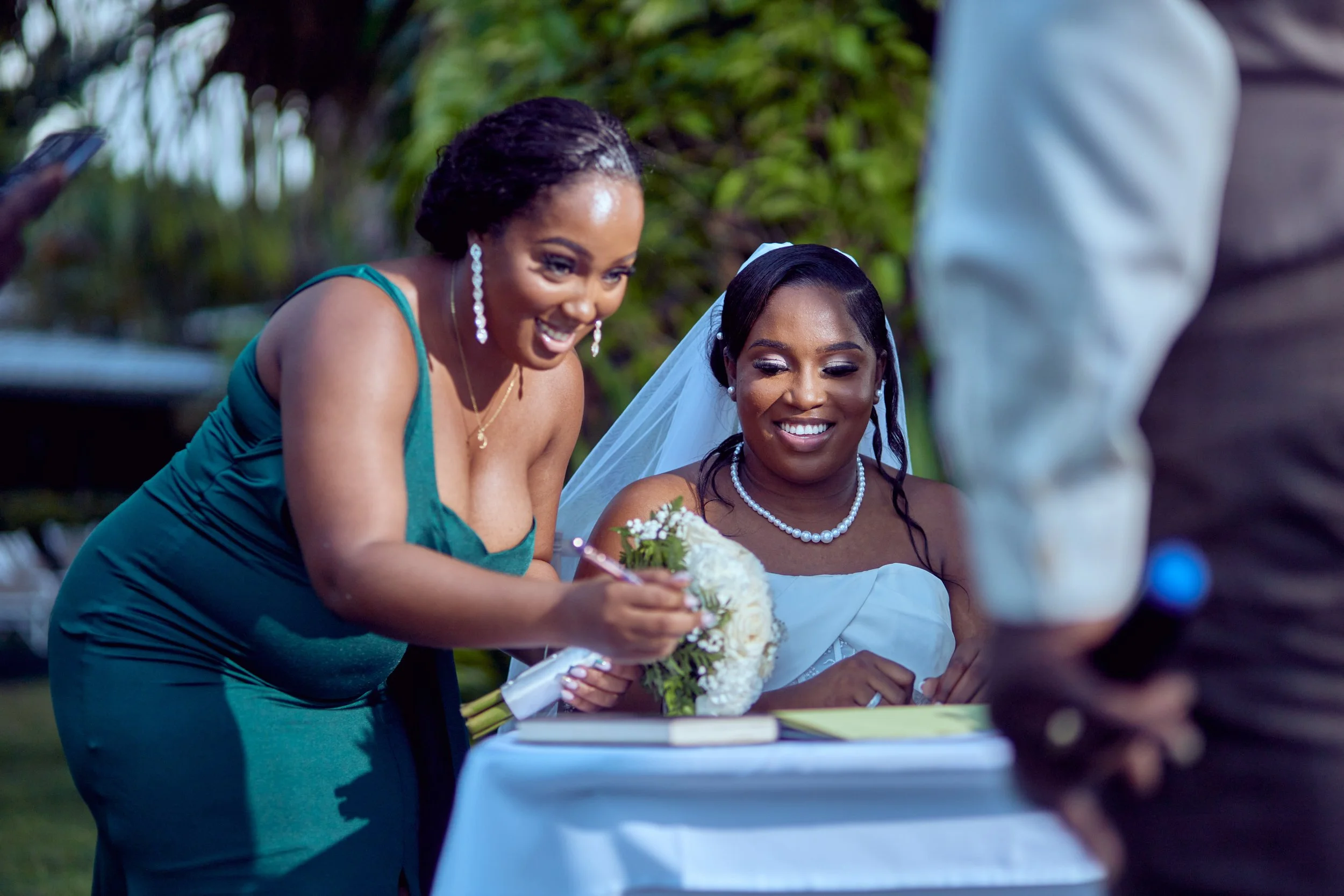 A woman in a teal dress signs a document at a wedding reception as a bride in a white dress and veil looks on and smiles.