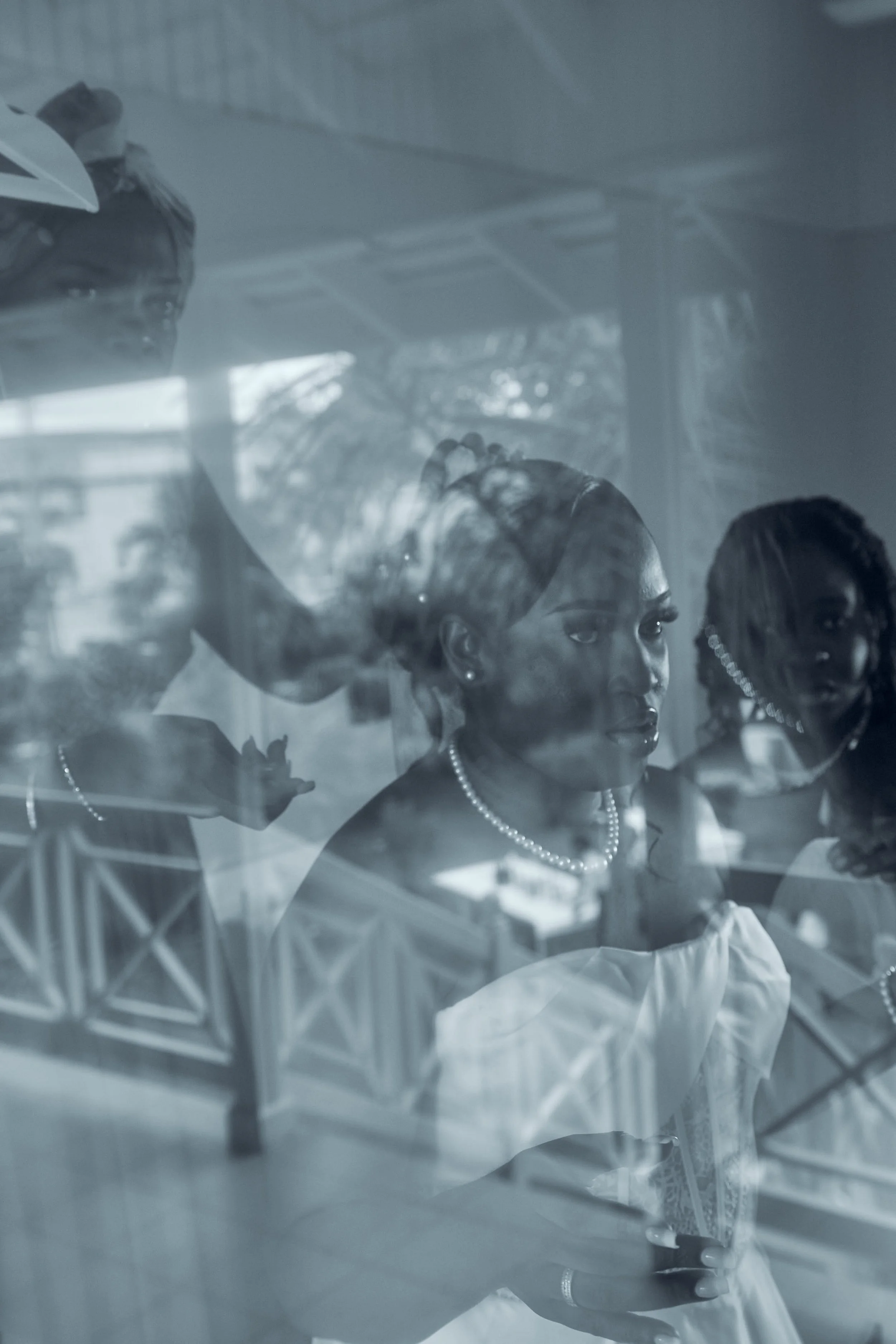 Reflection of two women with braided hair and pearl necklaces through a glass window, with one doing hair or makeup.
