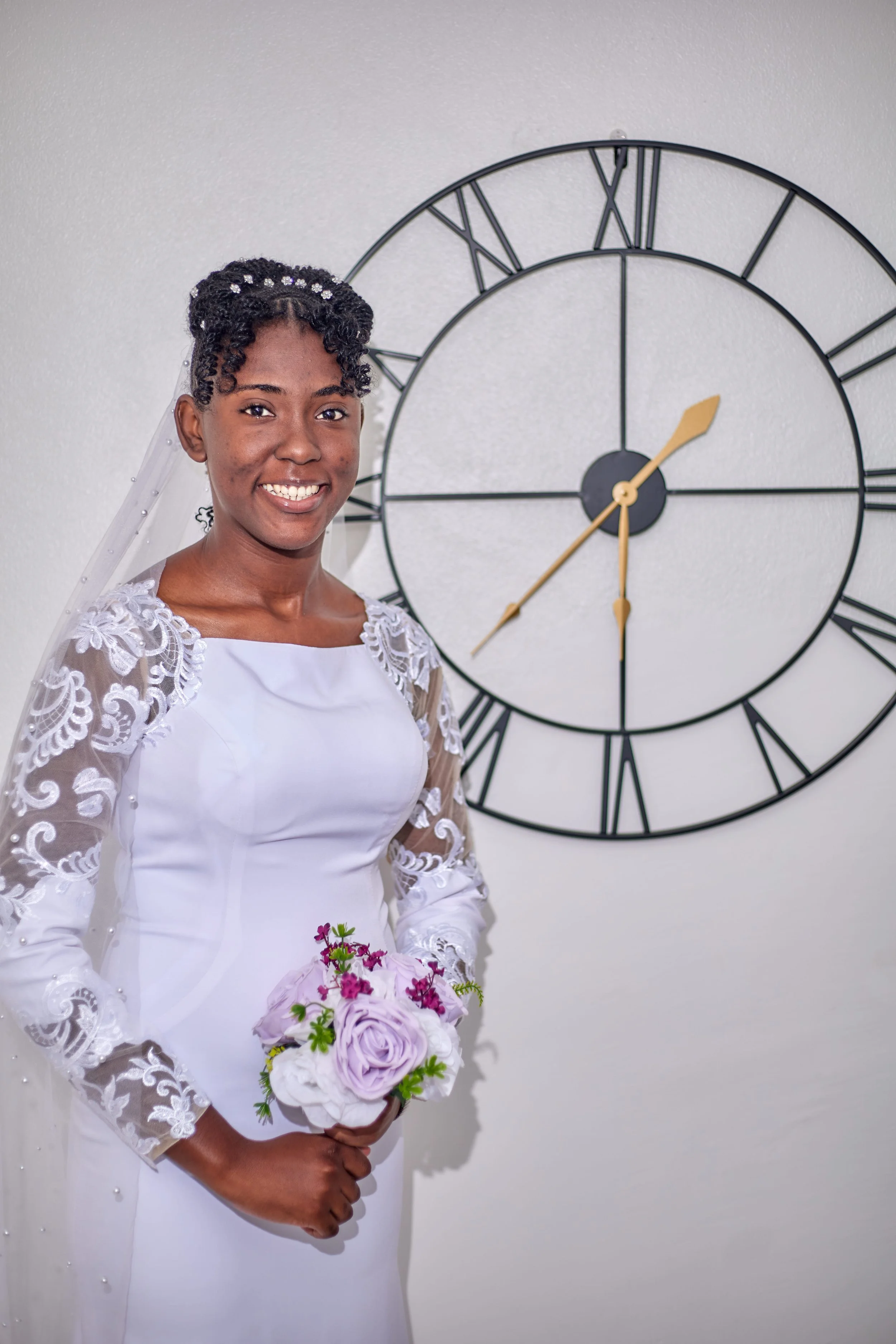 A bride in a white wedding dress with lace sleeves, holding a bouquet of lavender roses and pink flowers, standing in front of a large wall clock on a white wall.