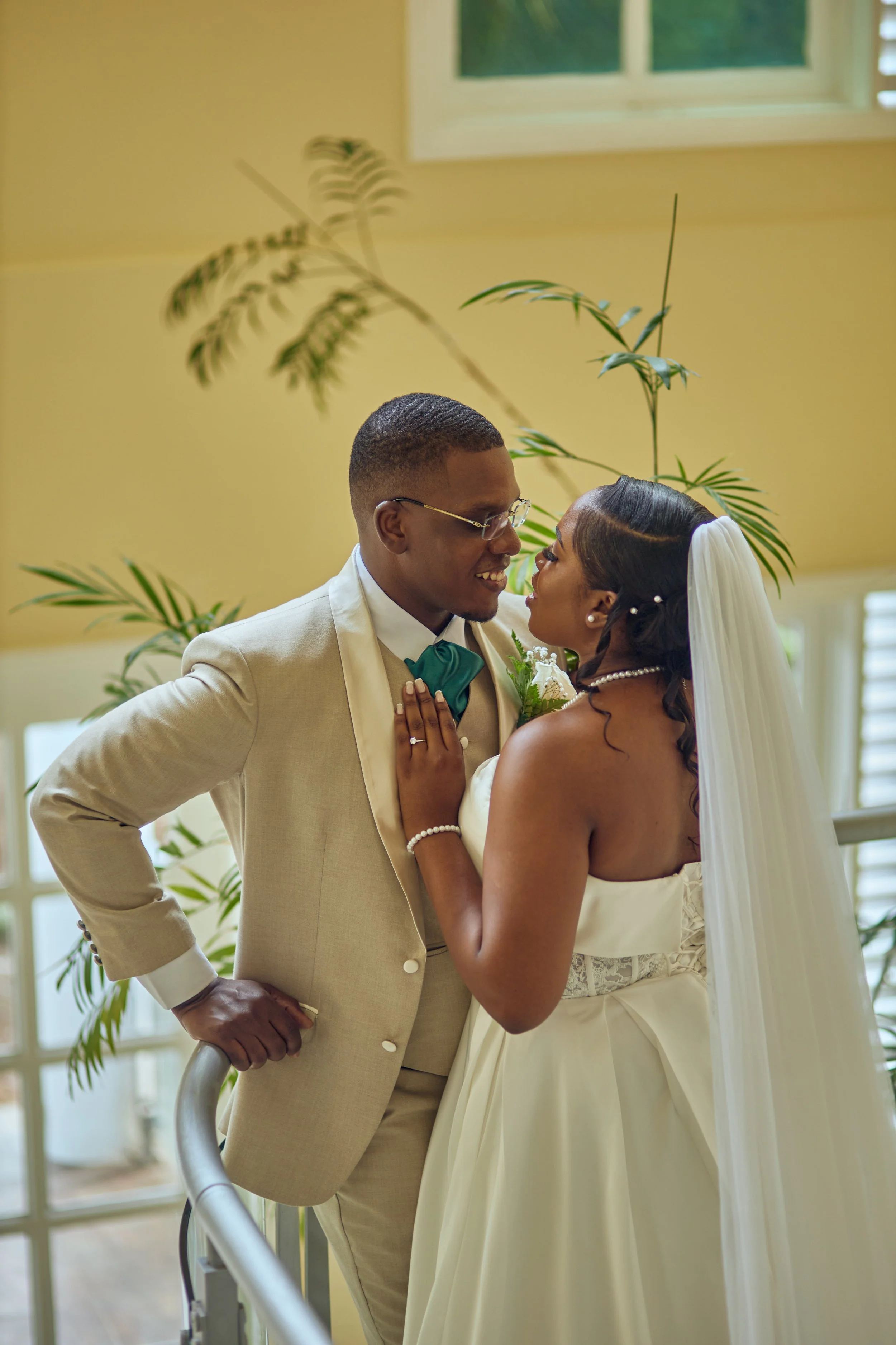 A newlywed couple sharing a loving moment indoors, with the groom wearing a beige suit and glasses, and the bride in a white strapless gown and veil, surrounded by green plants.