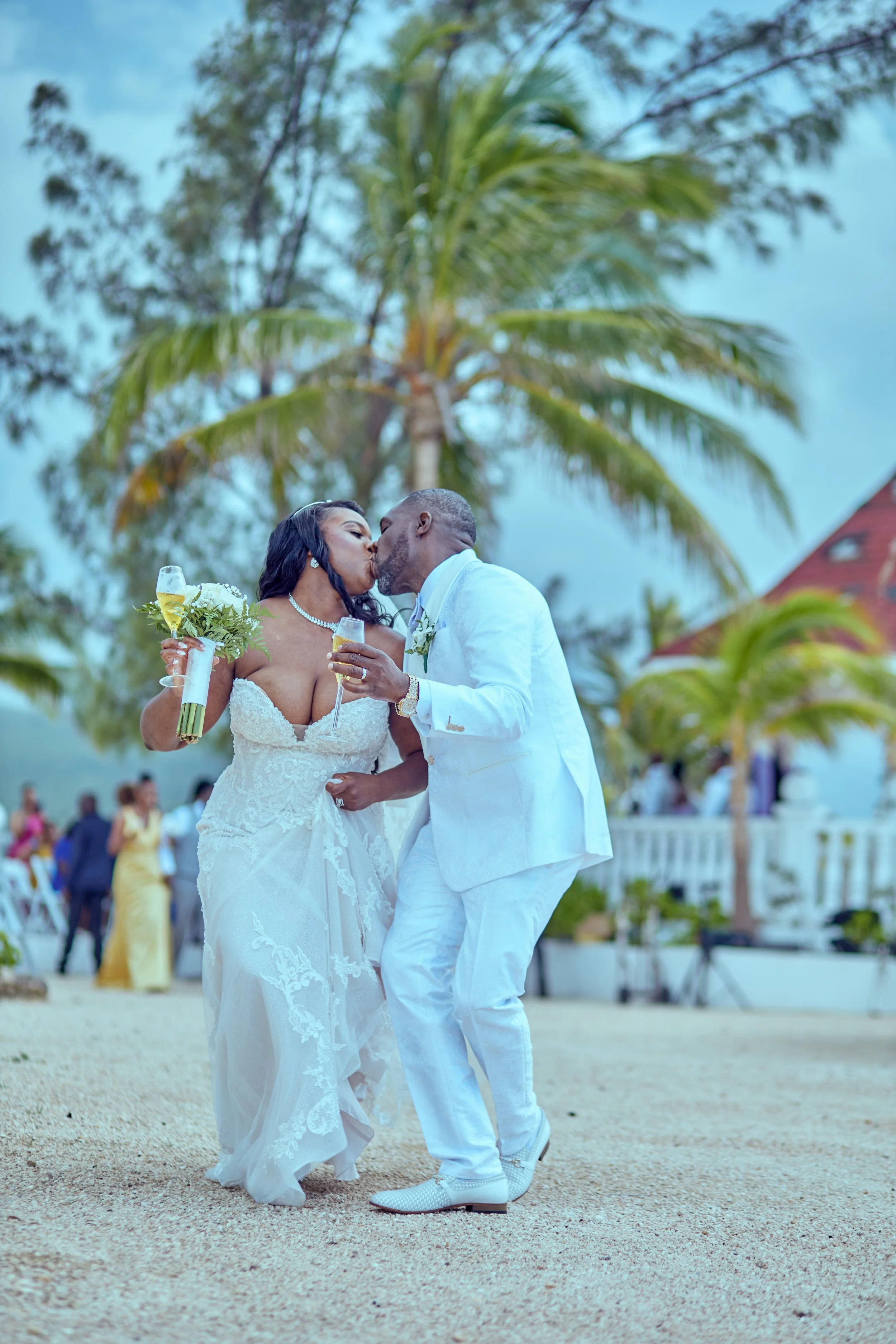 A newlywed couple sharing a kiss at their beach wedding ceremony, surrounded by palm trees and wedding guests.
