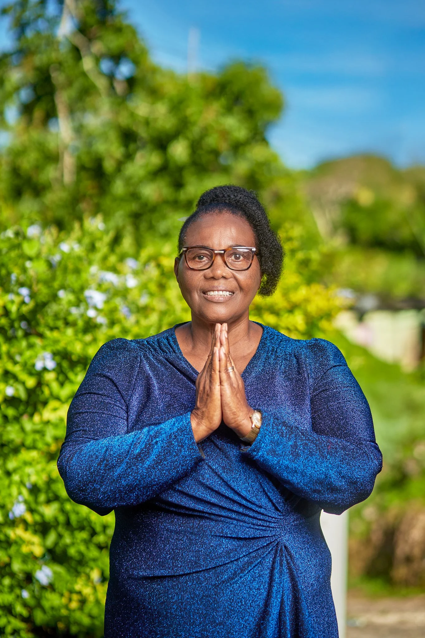 A woman with glasses and shoulder-length curly hair standing outdoors with hands pressed together in front of her chest, wearing a blue long-sleeve dress, surrounded by green trees and a blue sky.