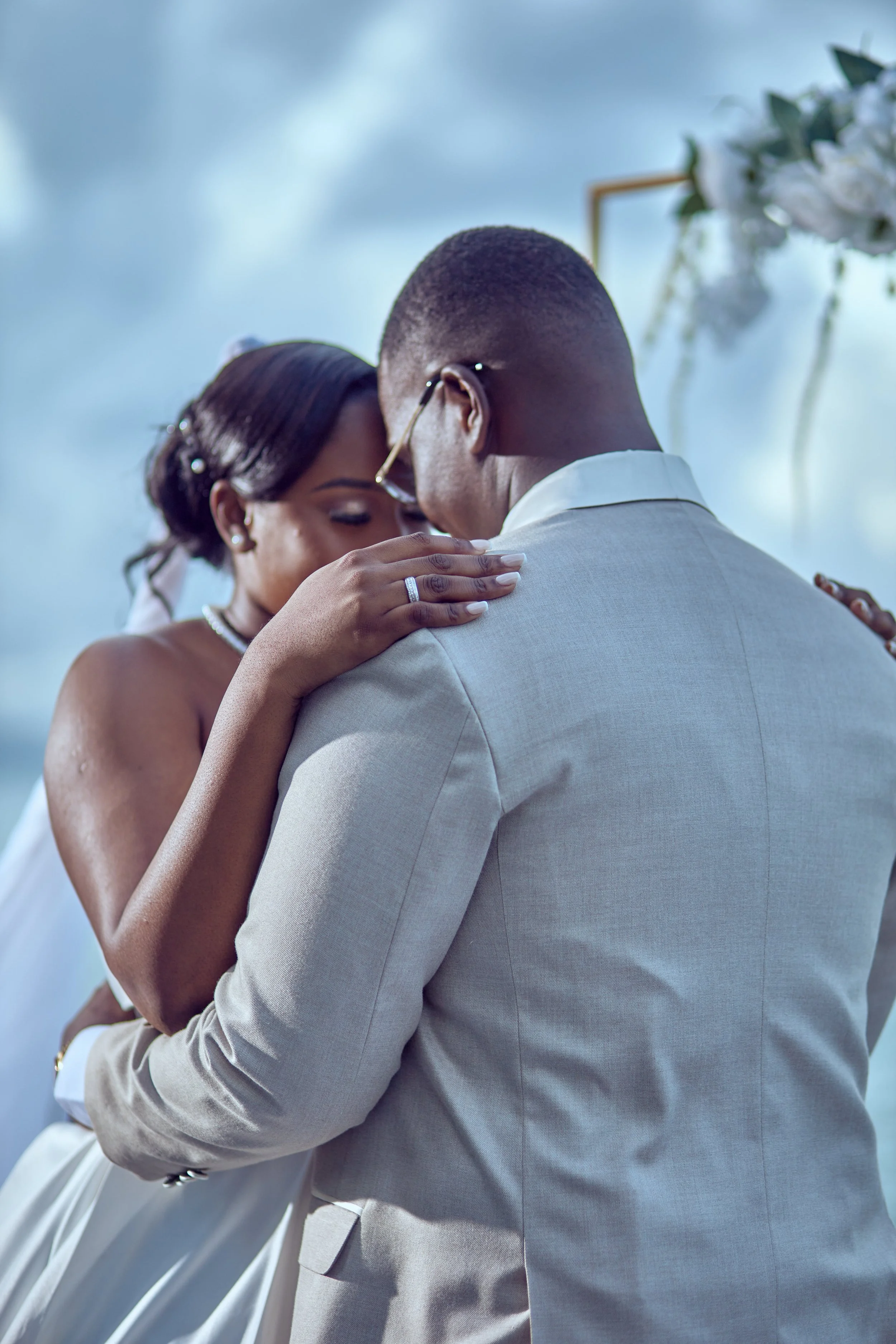 A bride and groom sharing a moment during their wedding ceremony, embracing closely with their foreheads touching against a cloudy sky backdrop.