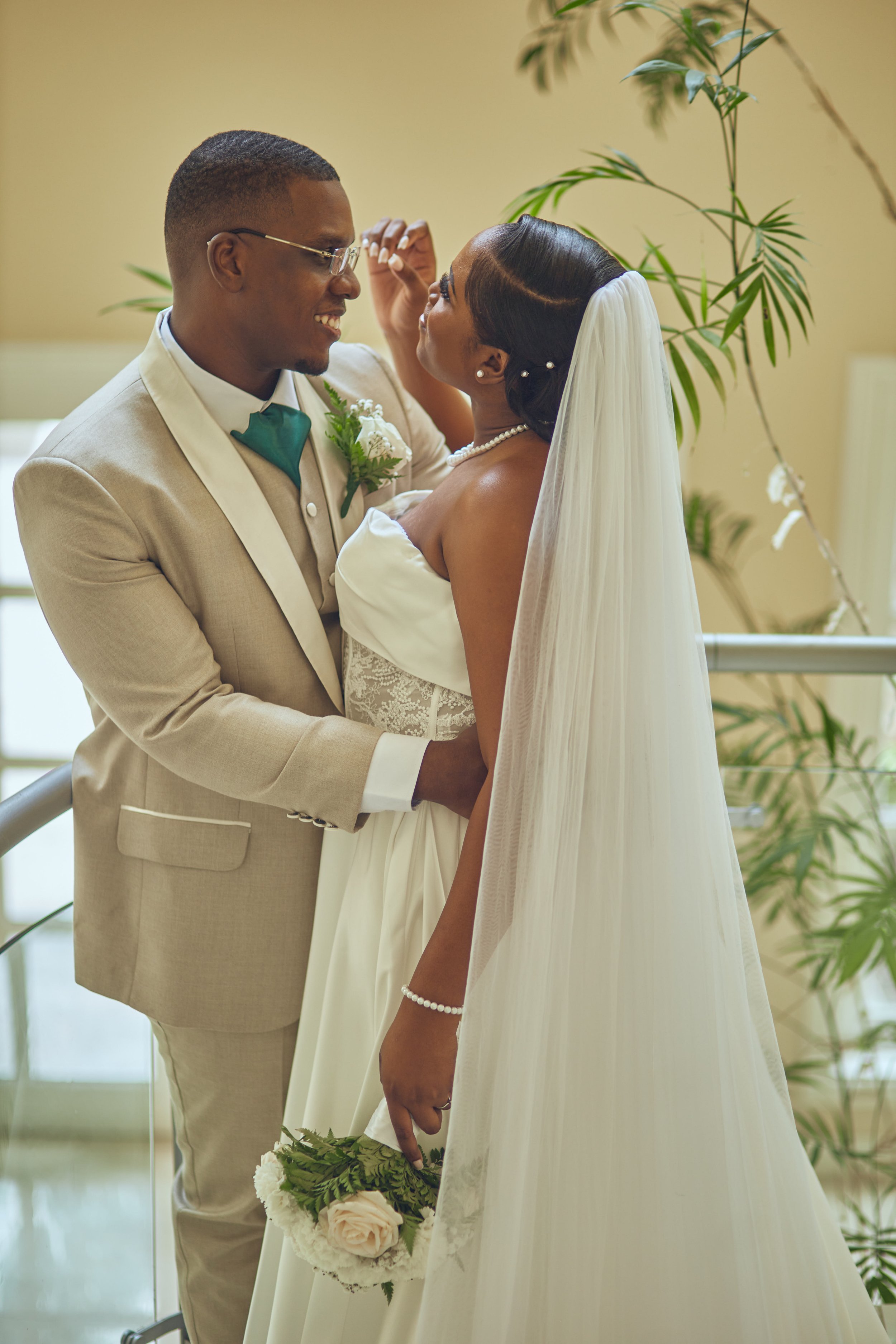 A wedding couple gazing at each other, the groom in a beige suit and green bowtie, and the bride in a strapless wedding gown with a veil, holding a bouquet of pink roses and greenery.