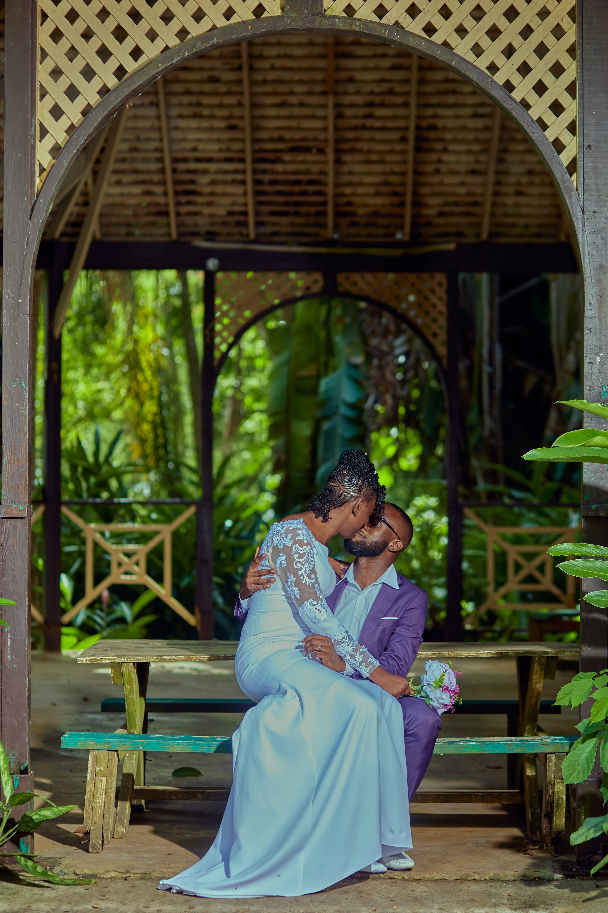 A couple dressed in wedding attire sharing a kiss while seated on a bench in a lush garden gazebo.