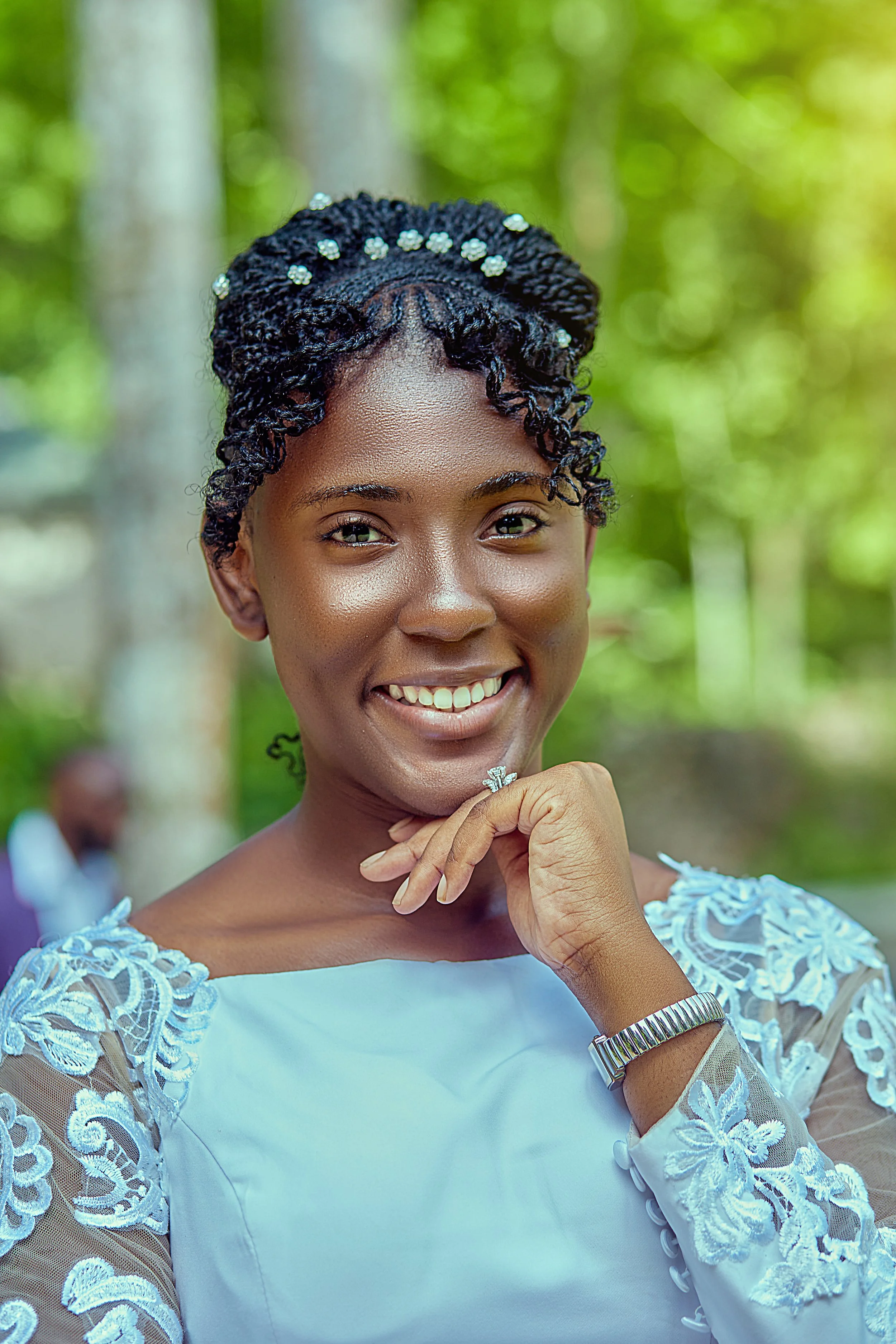 A smiling woman with styled black hair with decorative beads, wearing a white dress with lace details, a watch, a ring, and jewelry, outdoors with green foliage in the background.