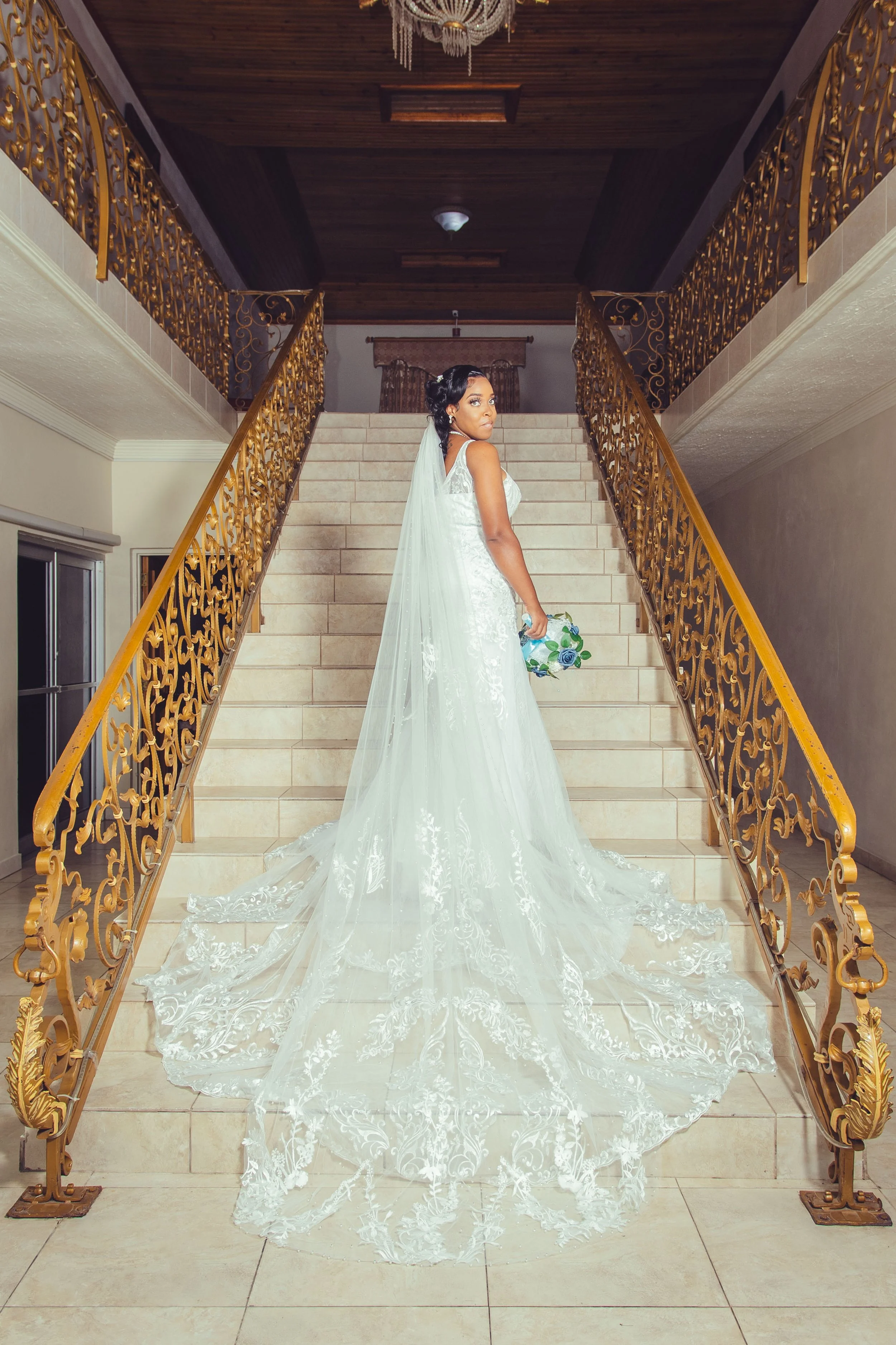 Bride standing at the top of a staircase in an elegant wedding dress with a long train and veil, holding a bouquet of flowers, inside a luxurious venue with gold ornate railings.