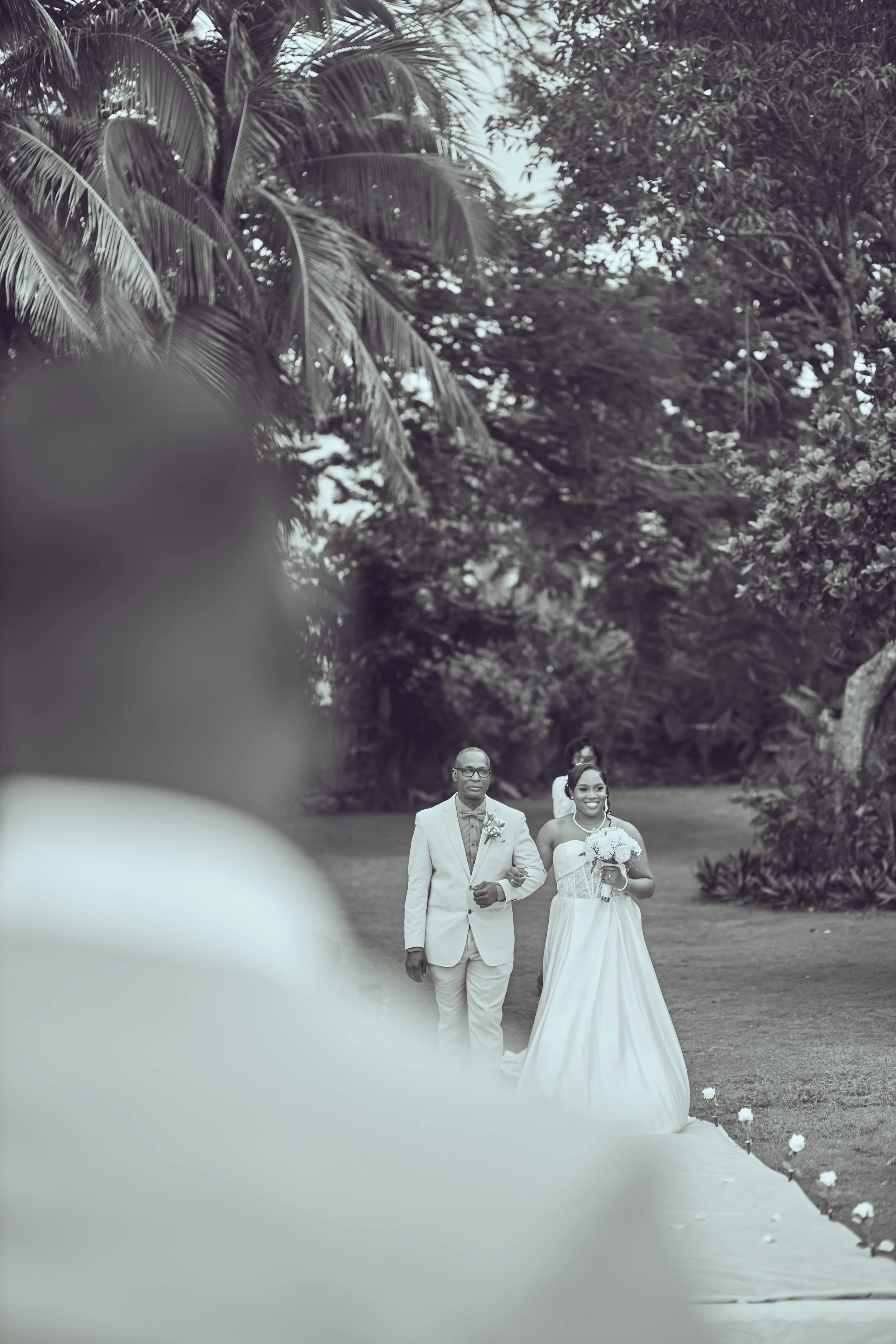 A bride and groom walking down an outdoor wedding aisle lined with flowers, surrounded by trees and foliage, with a blurred foreground.