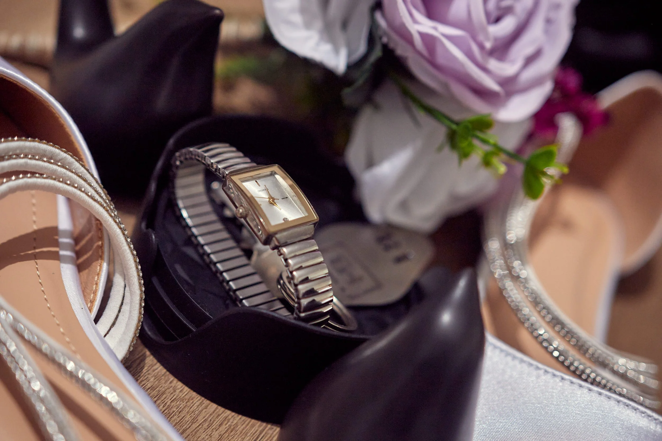 A collection of women's accessories, including a pair of beige high-heeled shoes, a silver watch, and a silver bracelet, arranged with a flower in a white wrapping.