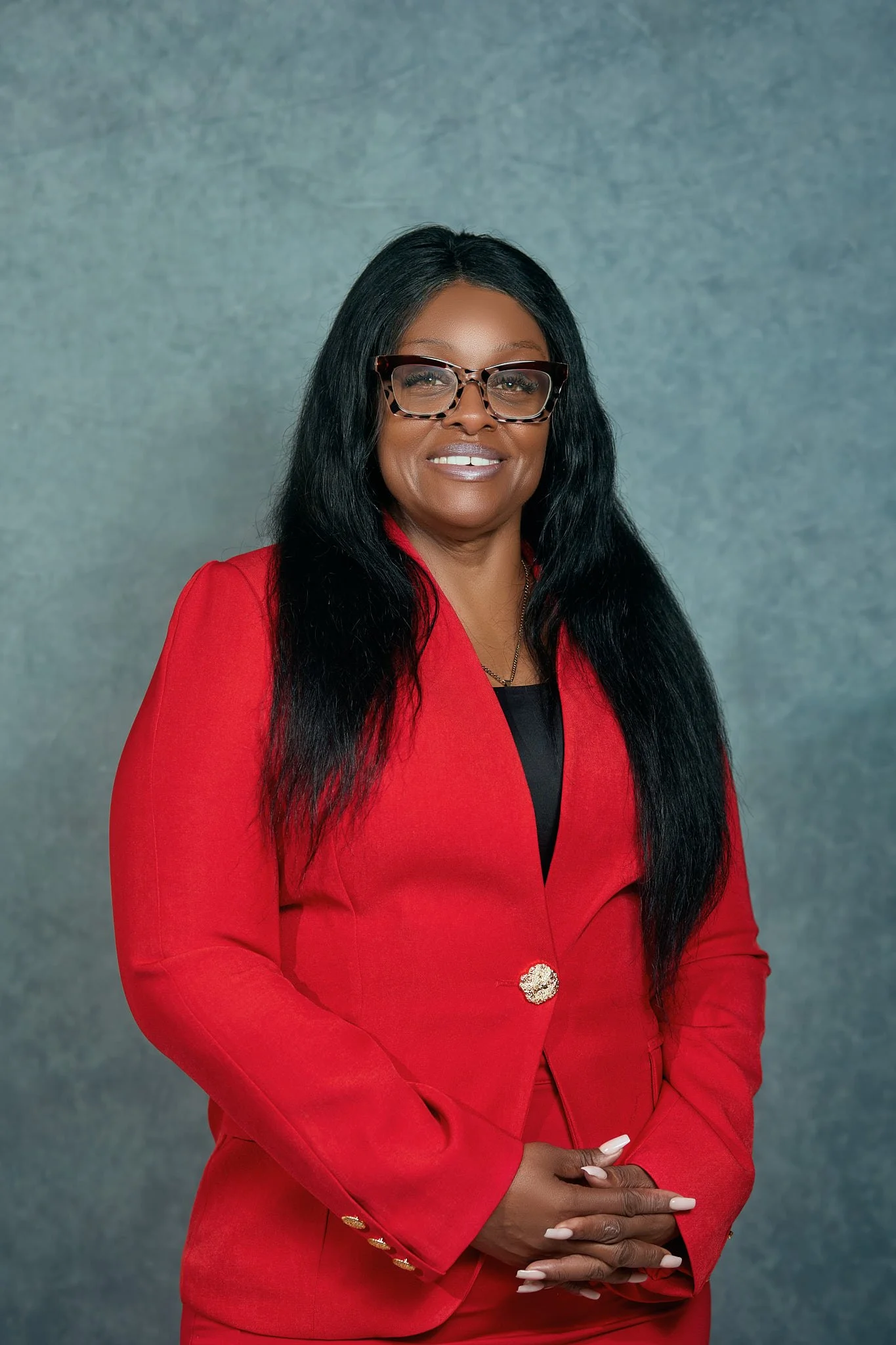 A woman with long black hair wearing glasses, a red blazer with gold buttons, and a black top, standing against a gray background.