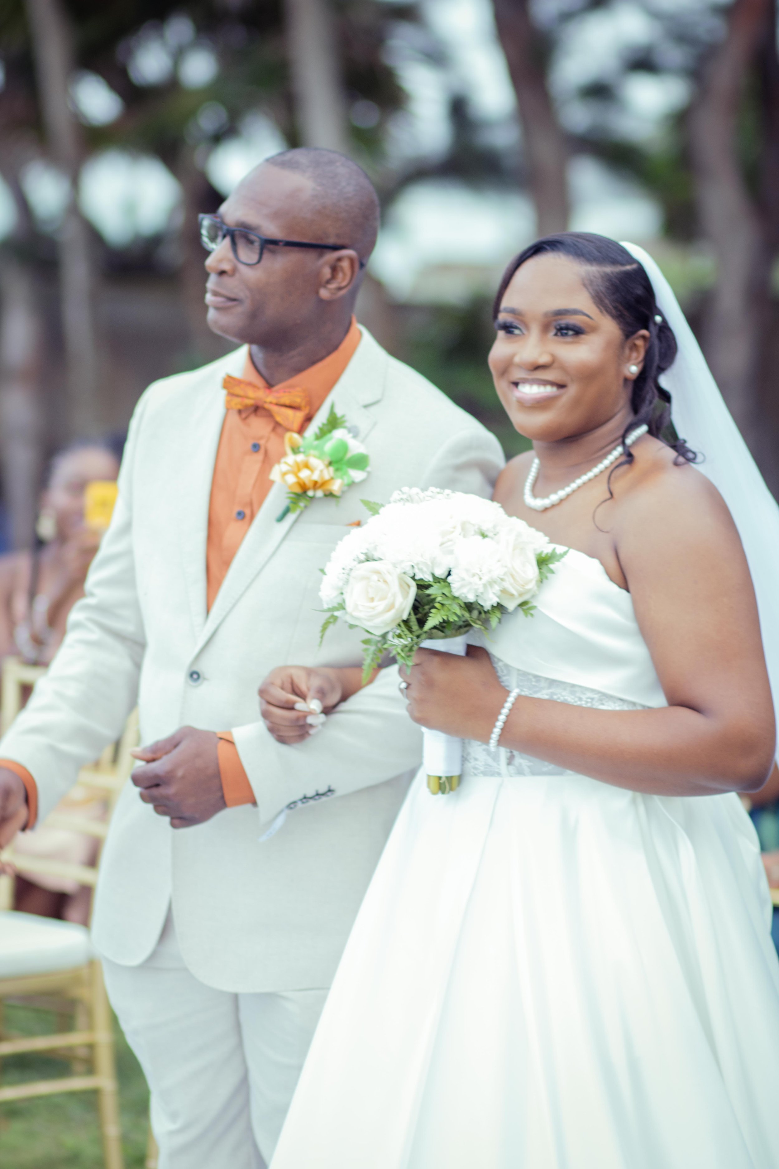A bride and an older man — possibly her father — walking down the aisle during a wedding ceremony outdoors. The bride is holding a bouquet of white flowers and is smiling, wearing a strapless white wedding gown with a pearl necklace and veil. The old