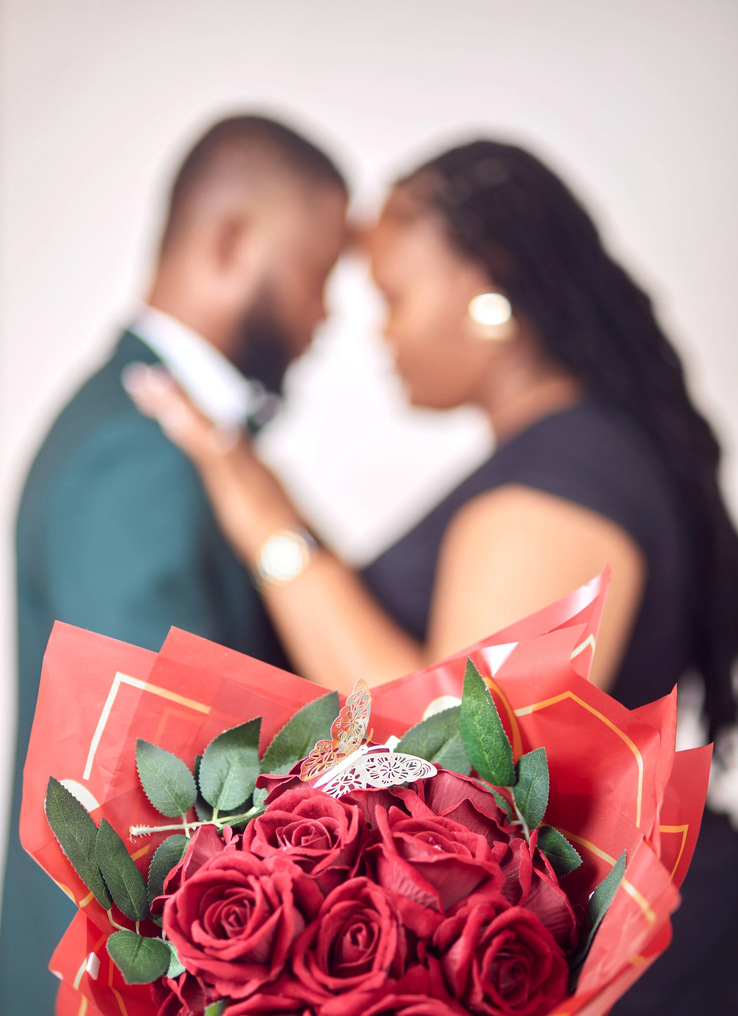 A blurred couple with foreheads touching and arms around each other in the background, with a bouquet of red roses and greenery in the foreground.