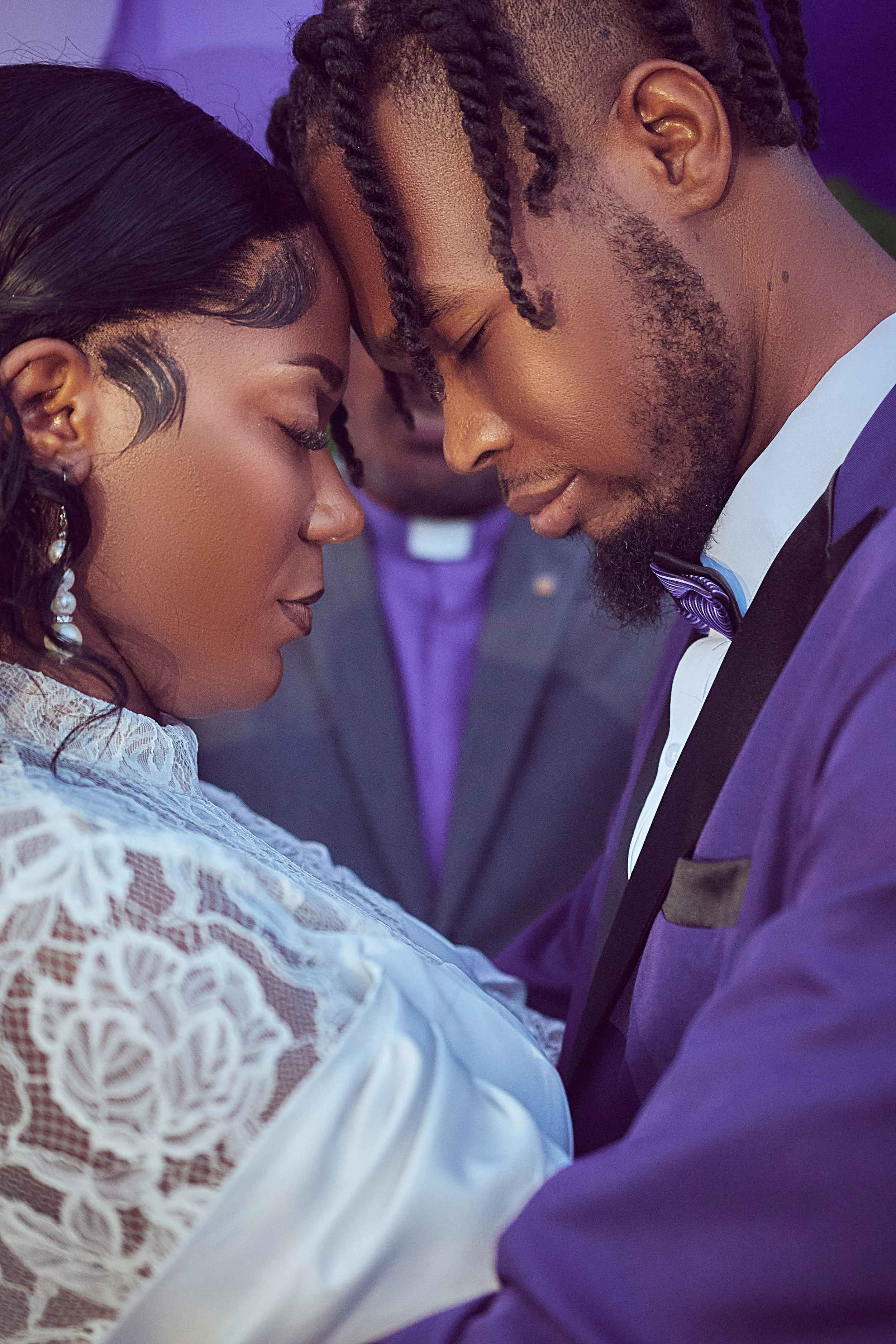 A bride and groom standing close with foreheads touching, eyes closed, during their wedding ceremony, with a person in the background.
