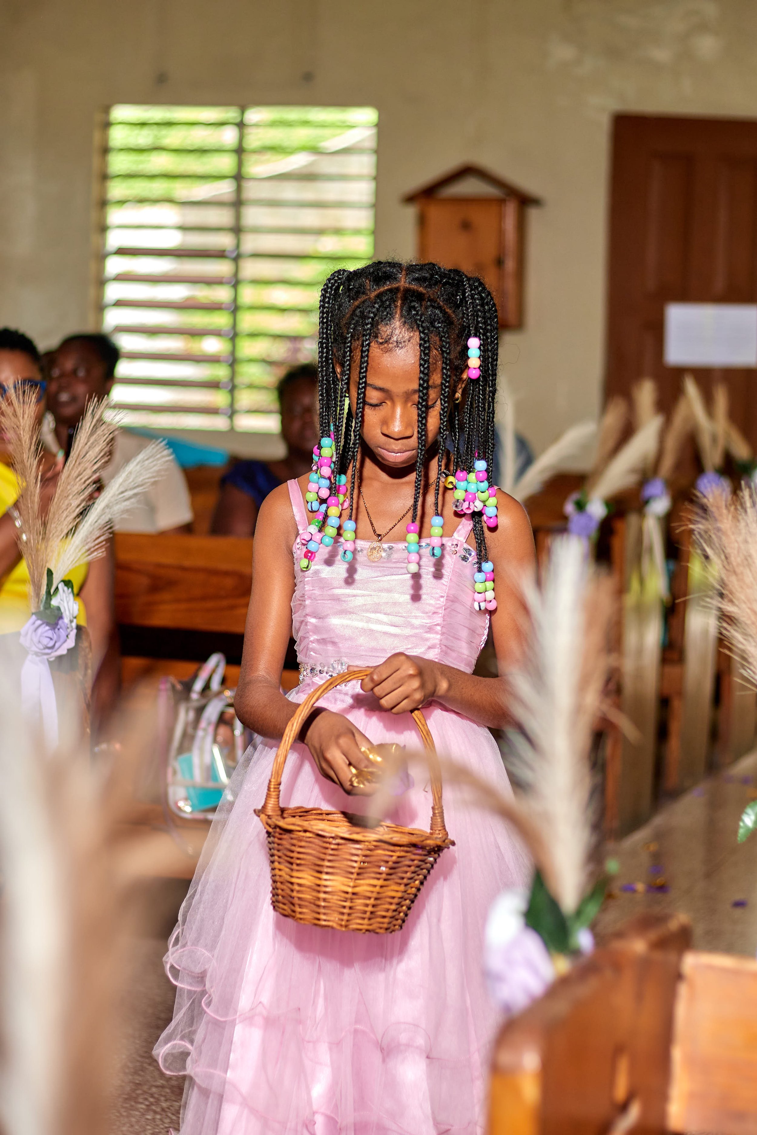 A young girl in a pink dress holding a small wicker basket in a decorated indoor setting, possibly during a church or community event, with other people in the background.