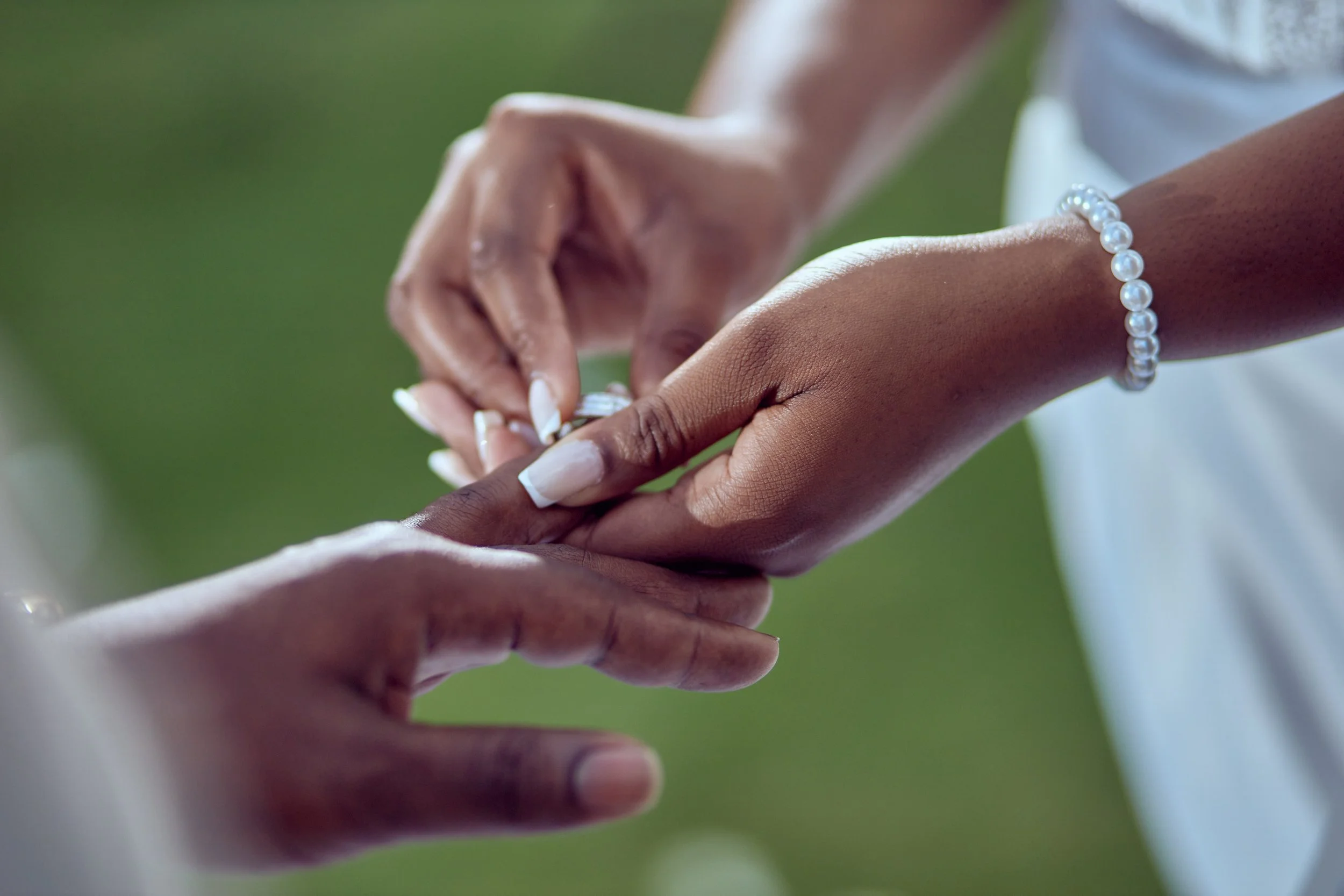 Close-up of a person placing a wedding ring on another person's finger during a wedding ceremony, with a pearl bracelet on the wrist.