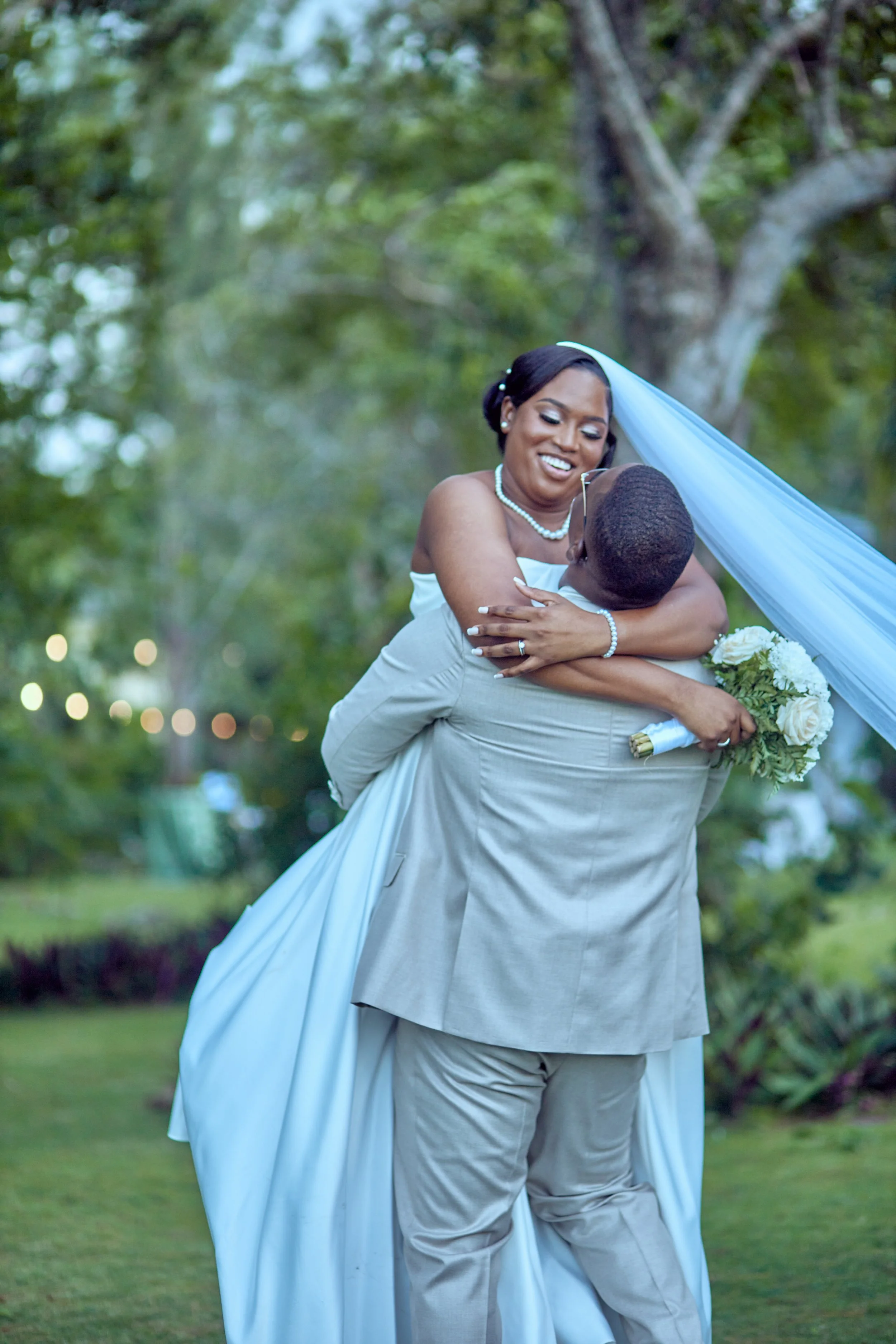 A bride is being lifted by a groom outdoors, holding a bouquet of white flowers, as they enjoy a joyful moment.