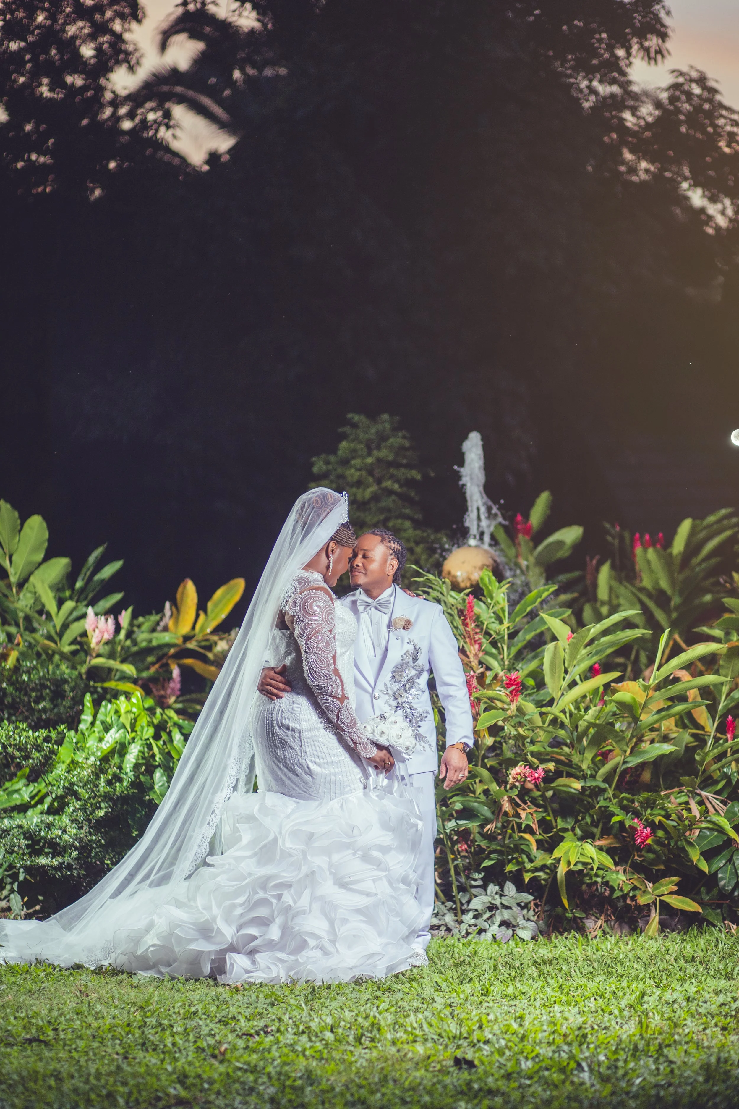 A bride and groom embrace during their wedding at night, surrounded by lush greenery and a fountain in the background.