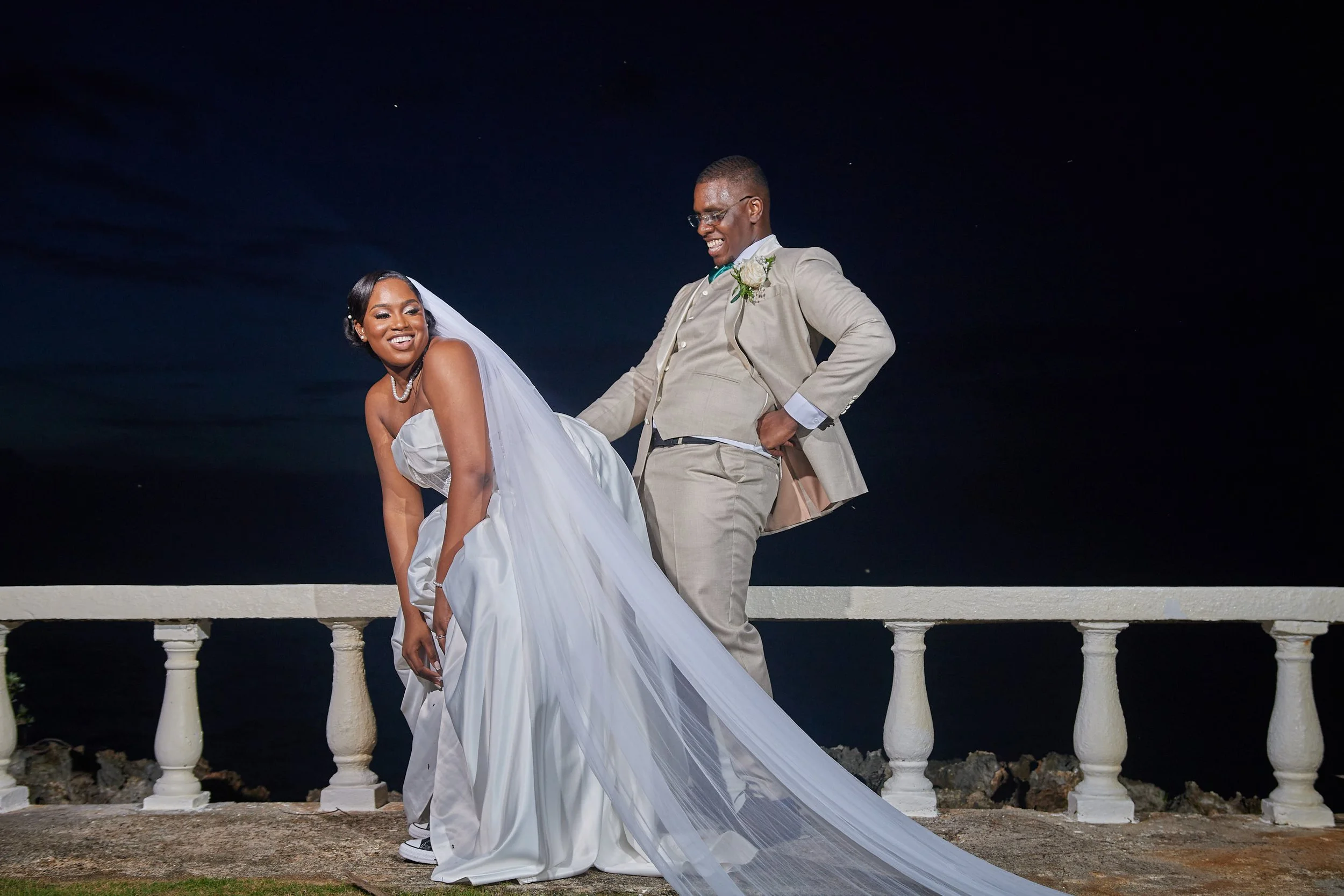 A joyful bride and groom at night on a terrace by the water, with the bride in a white wedding gown and veil, and the groom in a beige suit, sharing a playful moment.