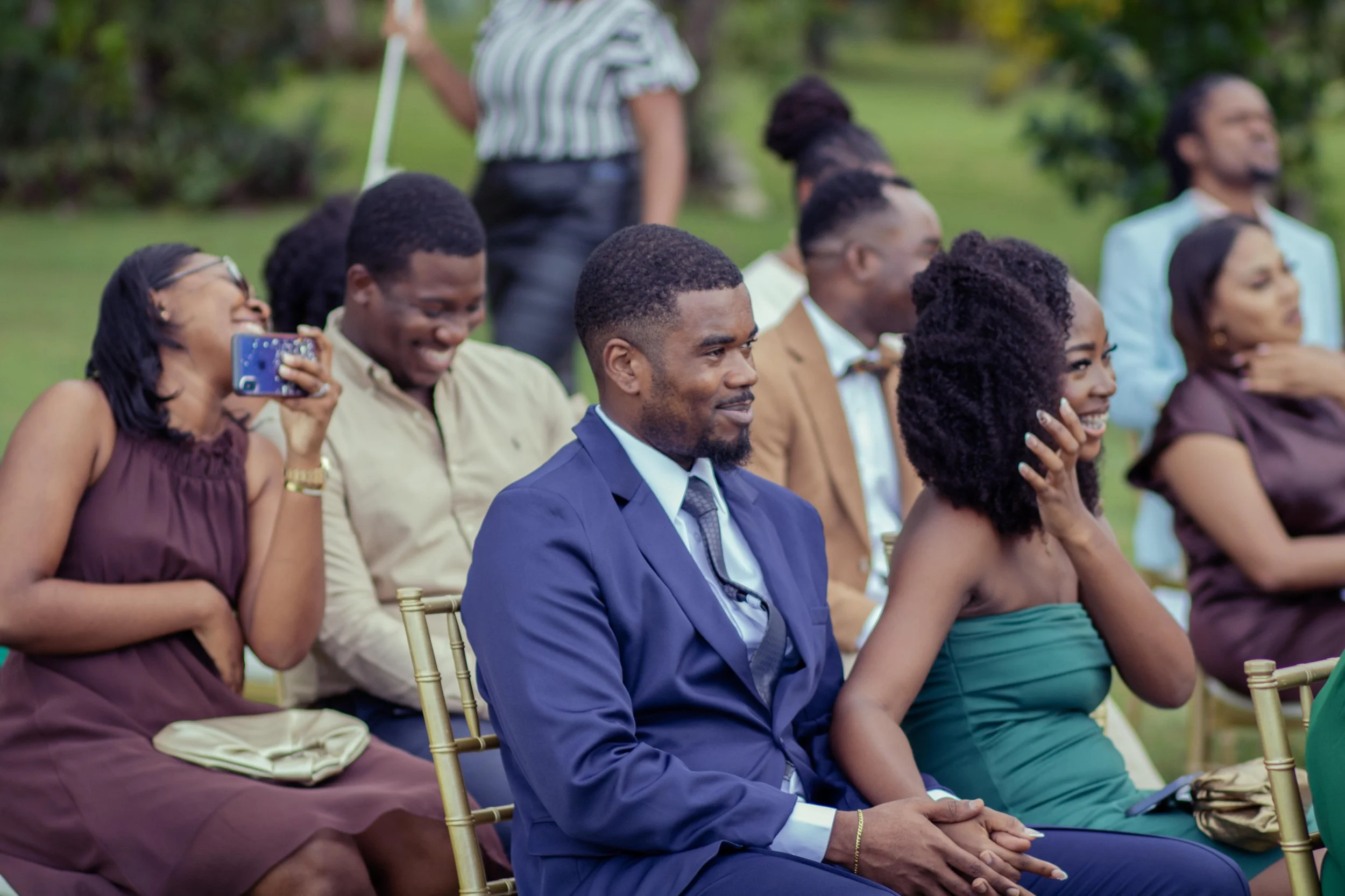 Group of people sitting outdoors at an event, smiling and laughing, dressed in formal attire.