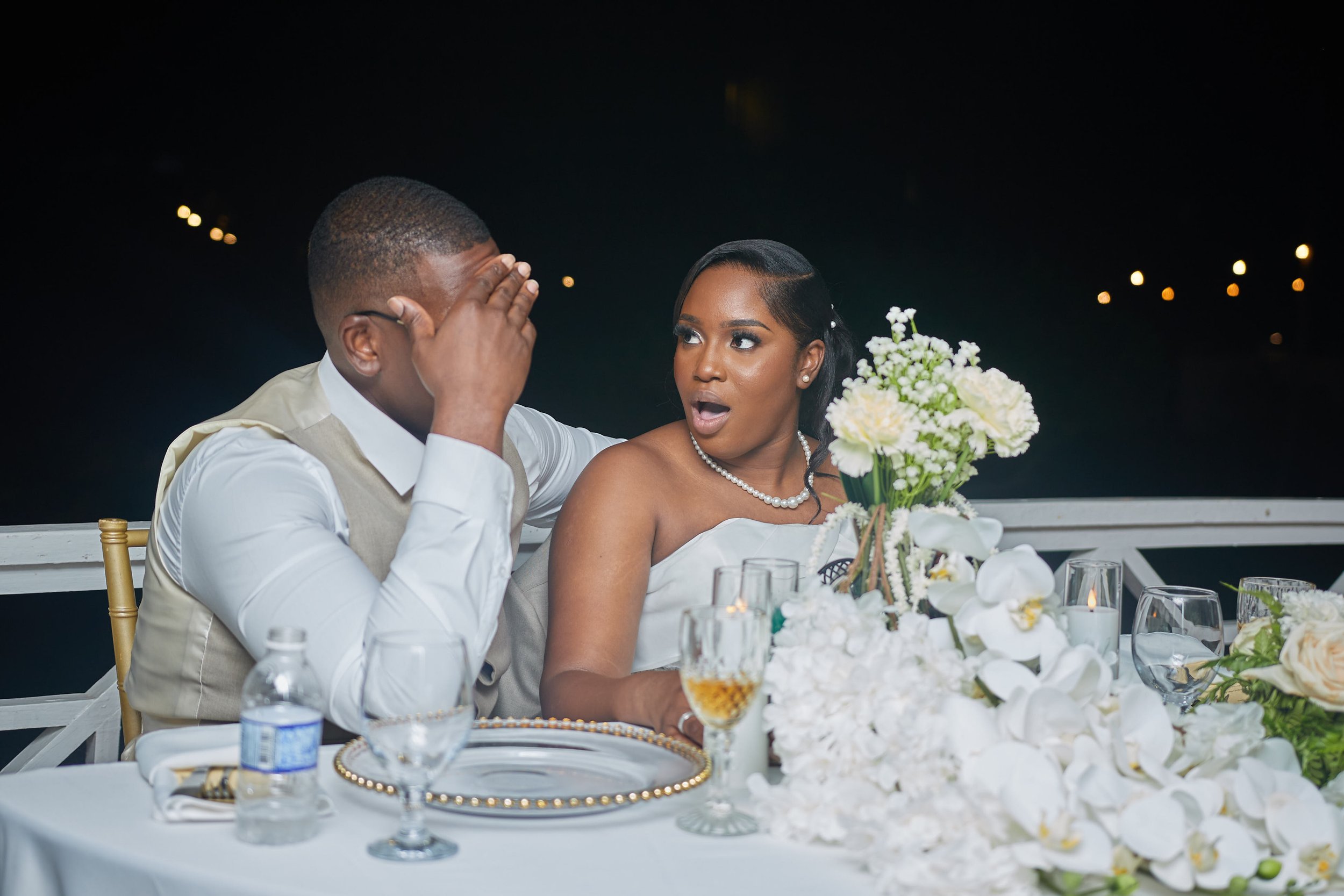 A bride and groom at a wedding reception, with the groom covering his face and the bride looking surprised, sitting at a decorated table with flowers, glasses, and a water bottle.